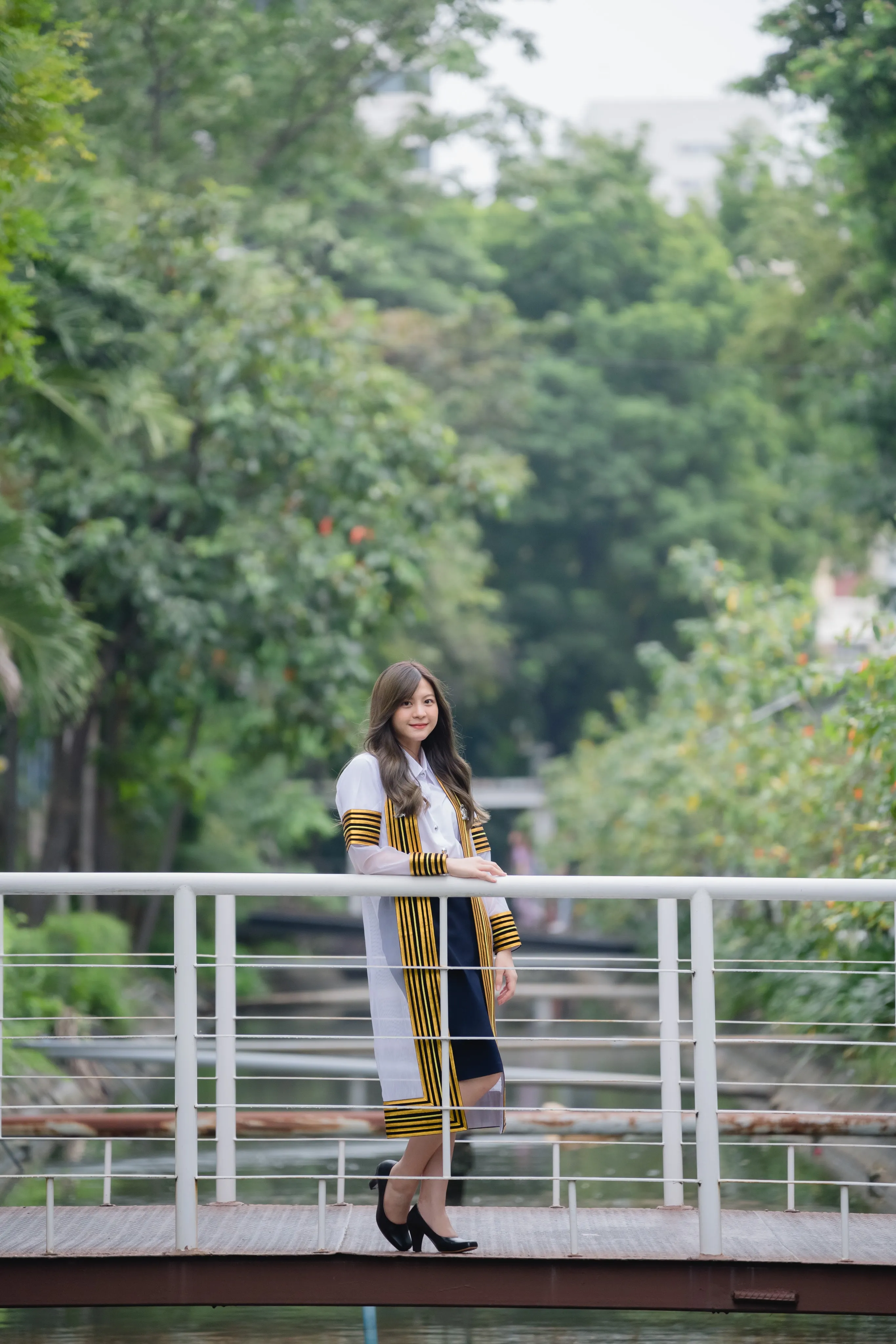 A graduate in a striped academic gown standing on a small footbridge surrounded by trees