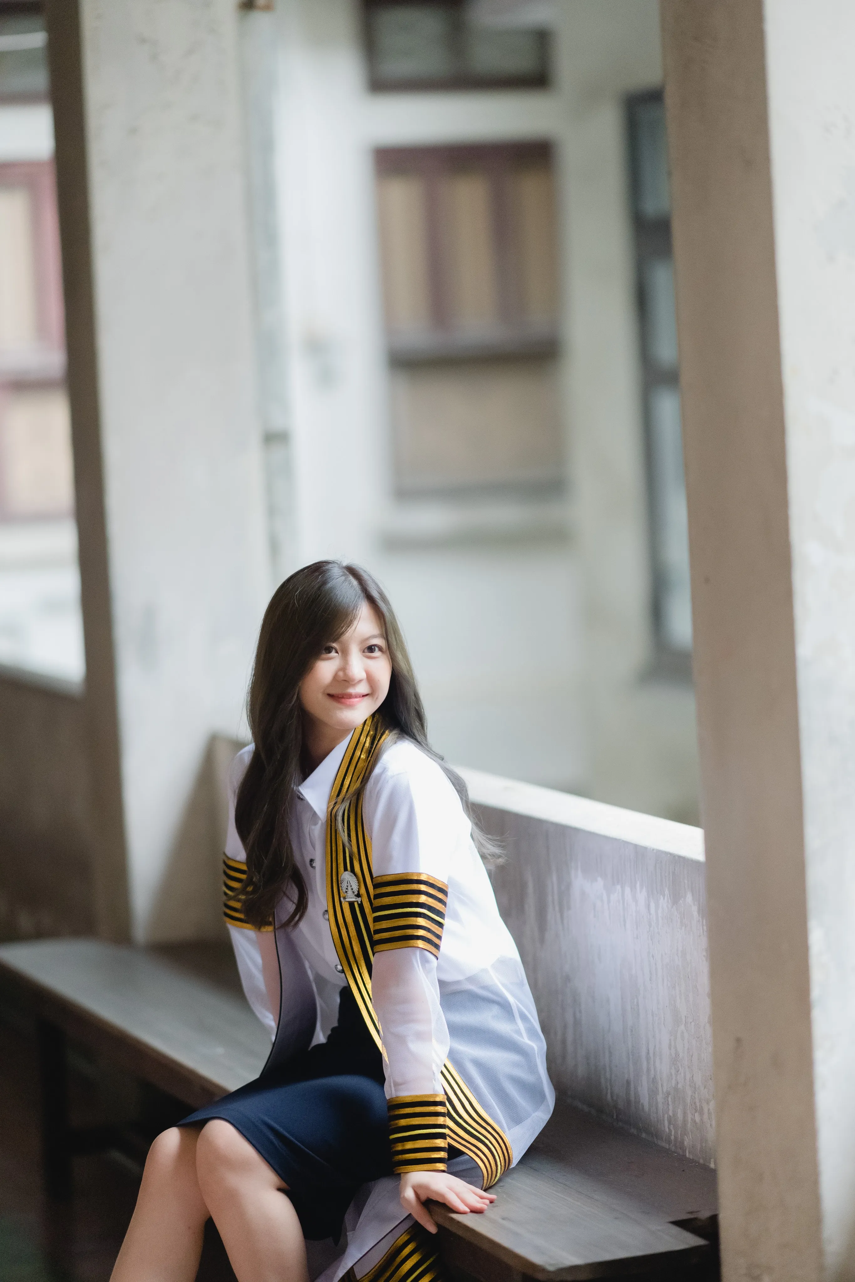 A smiling graduate in a striped academic gown sitting on a bench in a shaded campus walkway