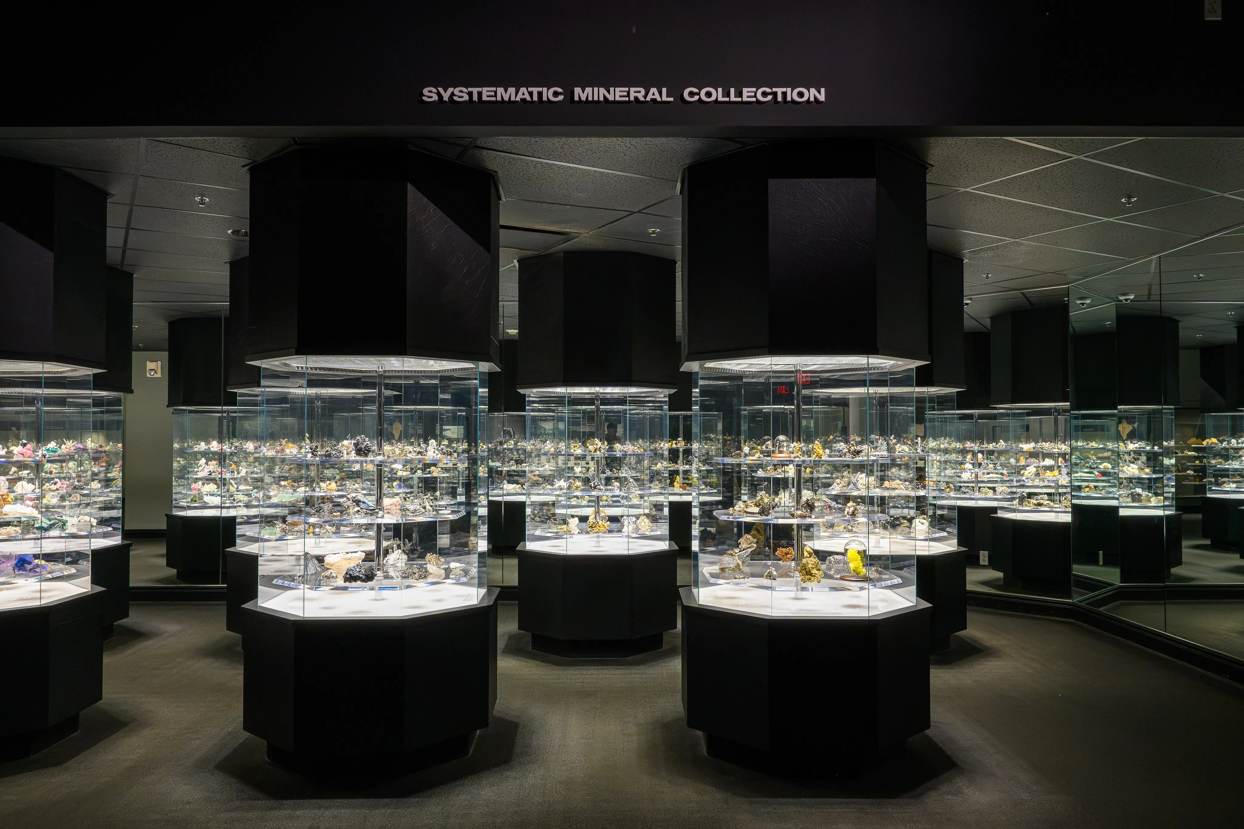 A dark gallery of illuminated display cases in the "Systematic Mineral Collection" exhibit.
