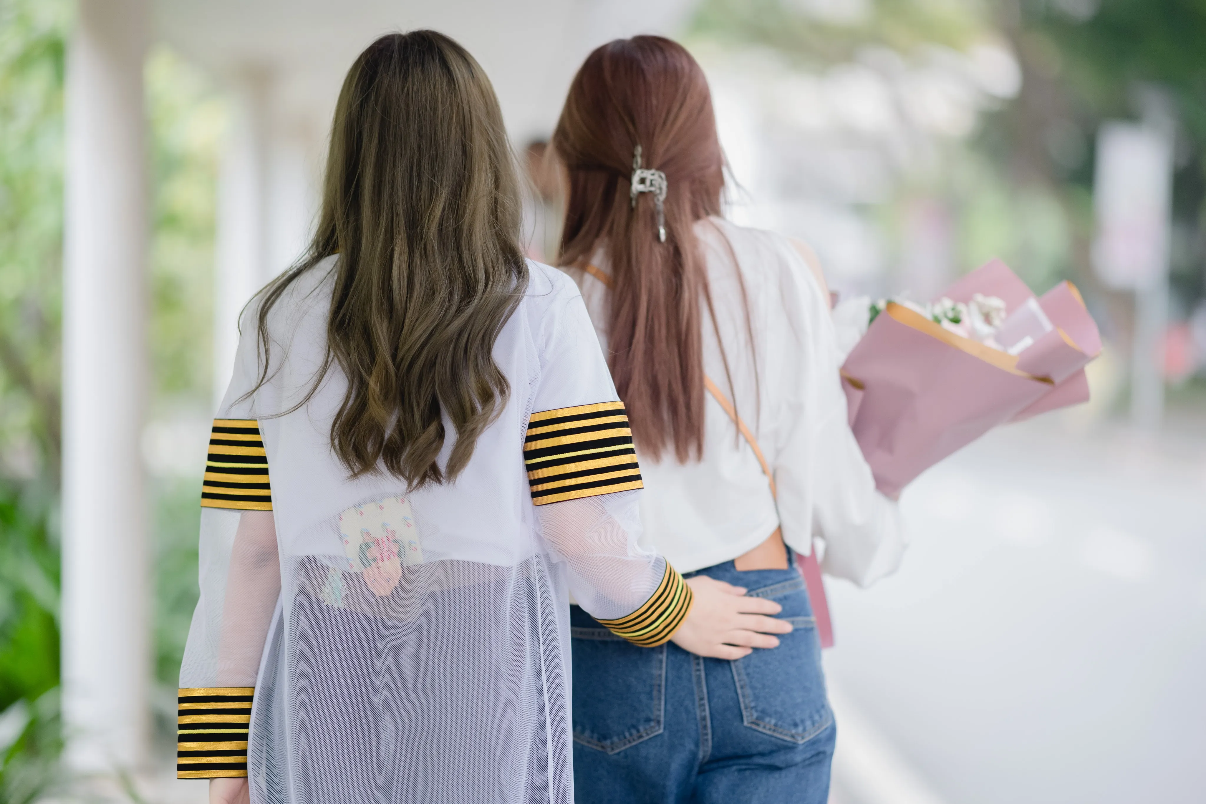 A graduate seen from behind with her arm around a friend carrying a bouquet