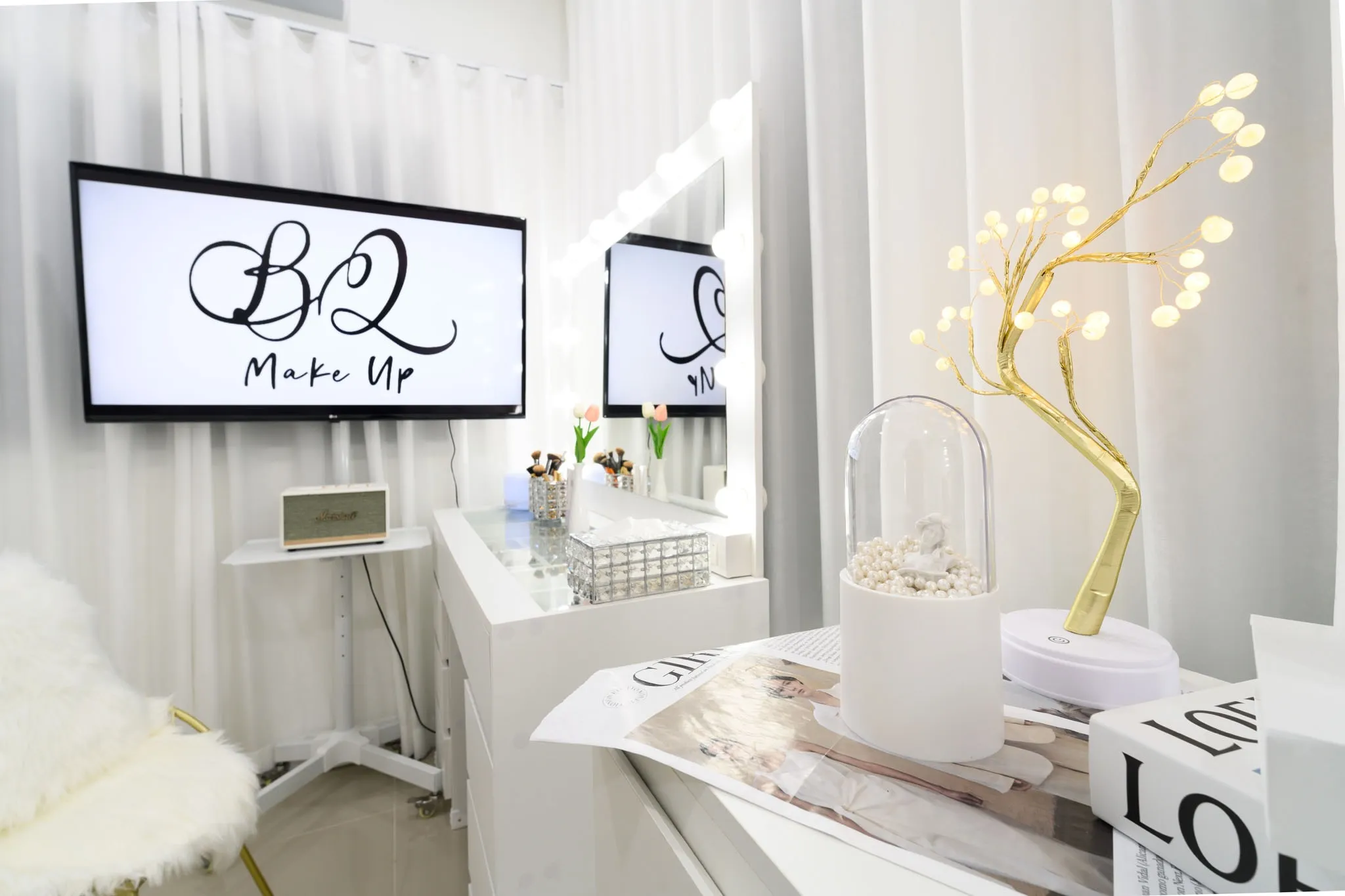 A white makeup station with a lit mirror, a golden tree lamp, and a screen showing the studio logo