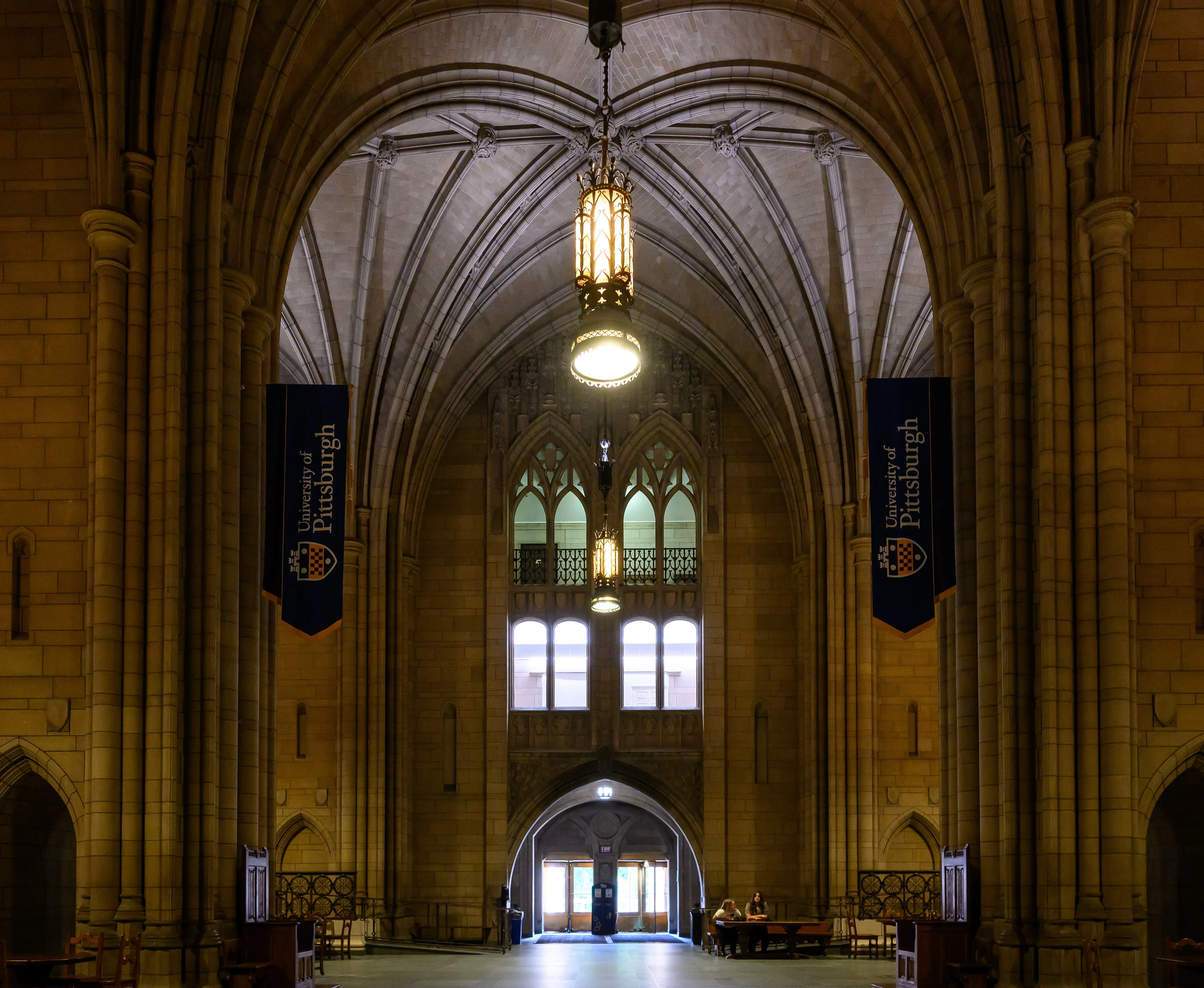 A vaulted Gothic hallway hung with university banners, leading toward an arched doorway.