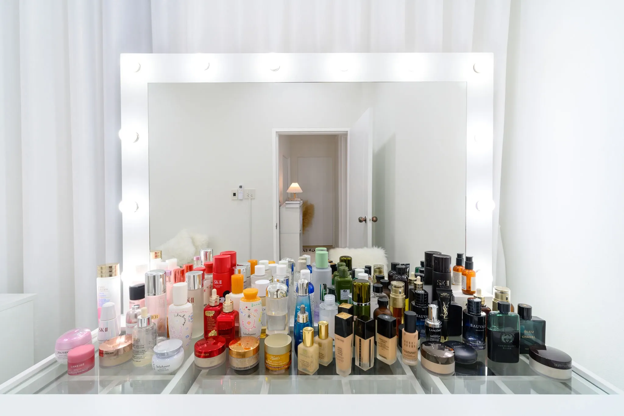A lit vanity mirror above a glass counter crowded with cosmetics and perfume bottles