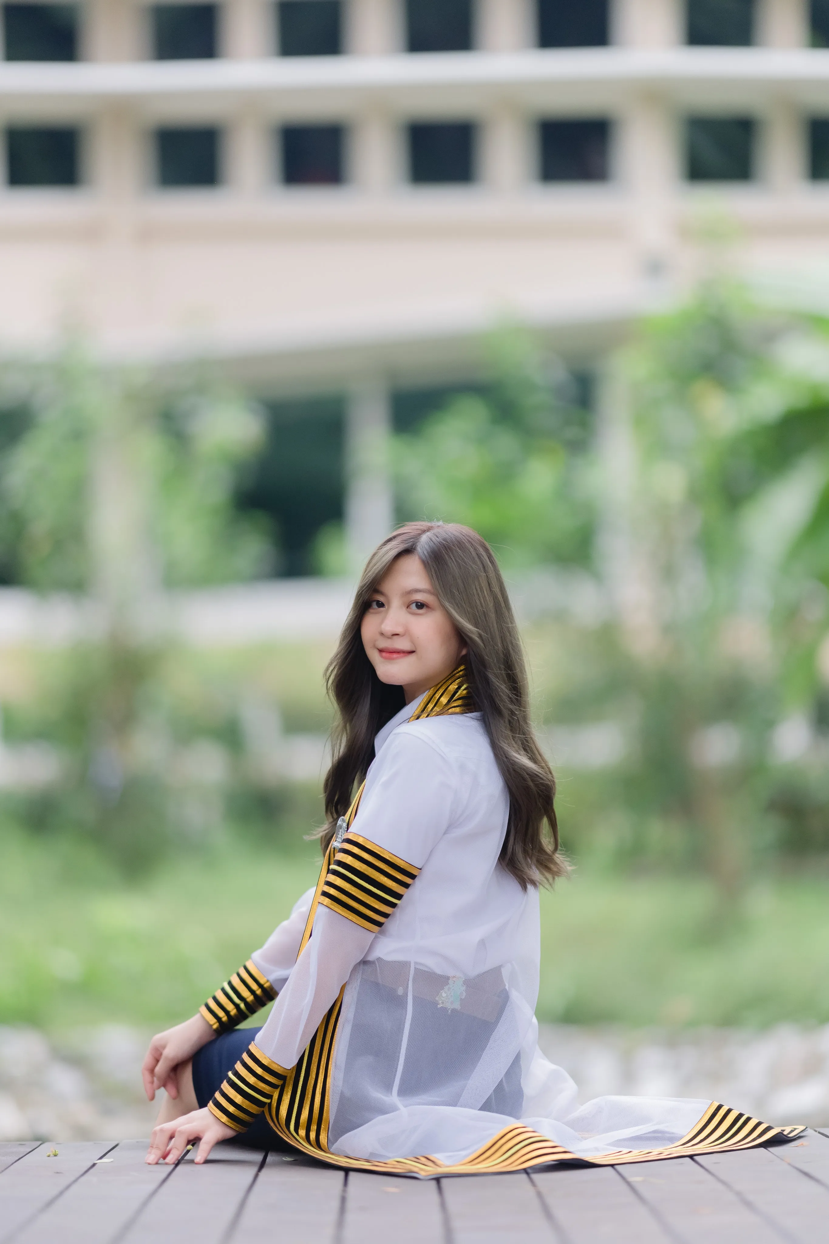 A graduate in a striped academic gown sitting on a wooden deck and smiling over her shoulder