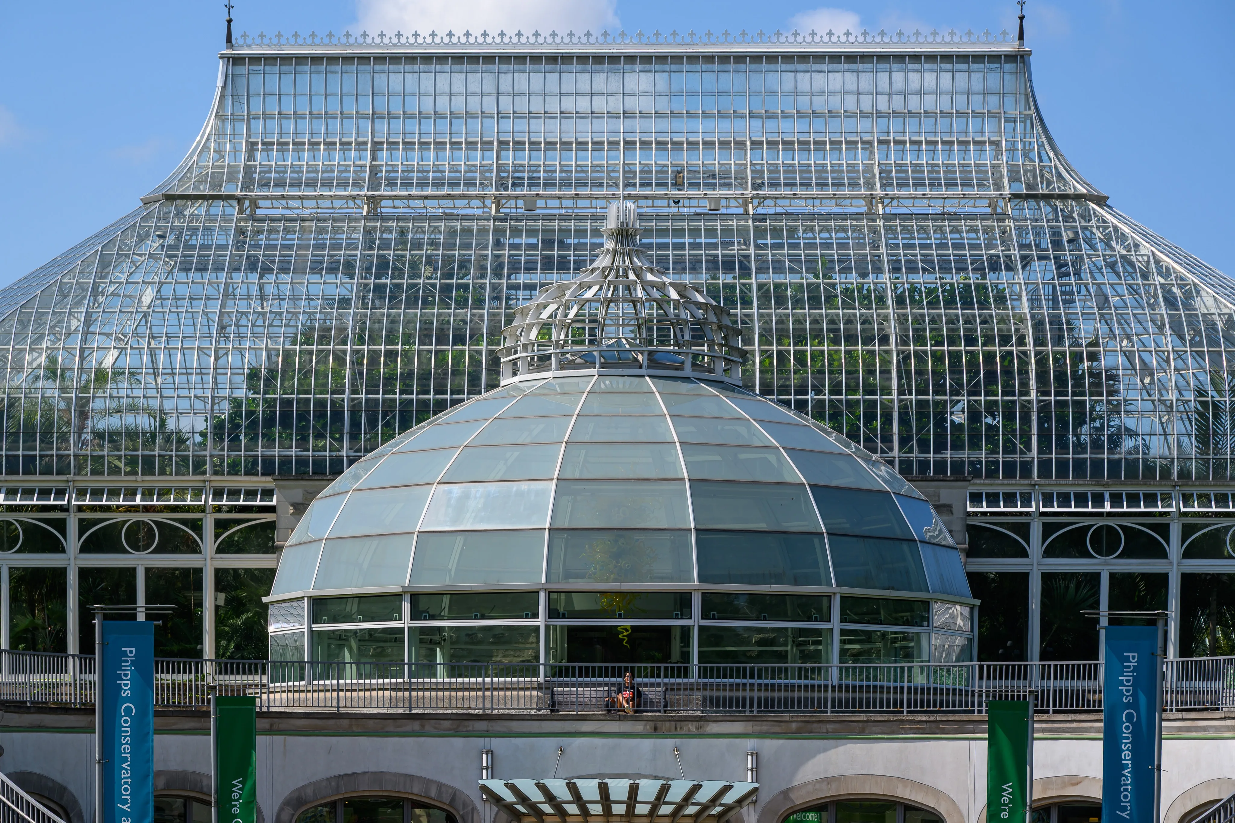 The glass dome and Victorian ironwork facade of a botanical conservatory against a blue sky.