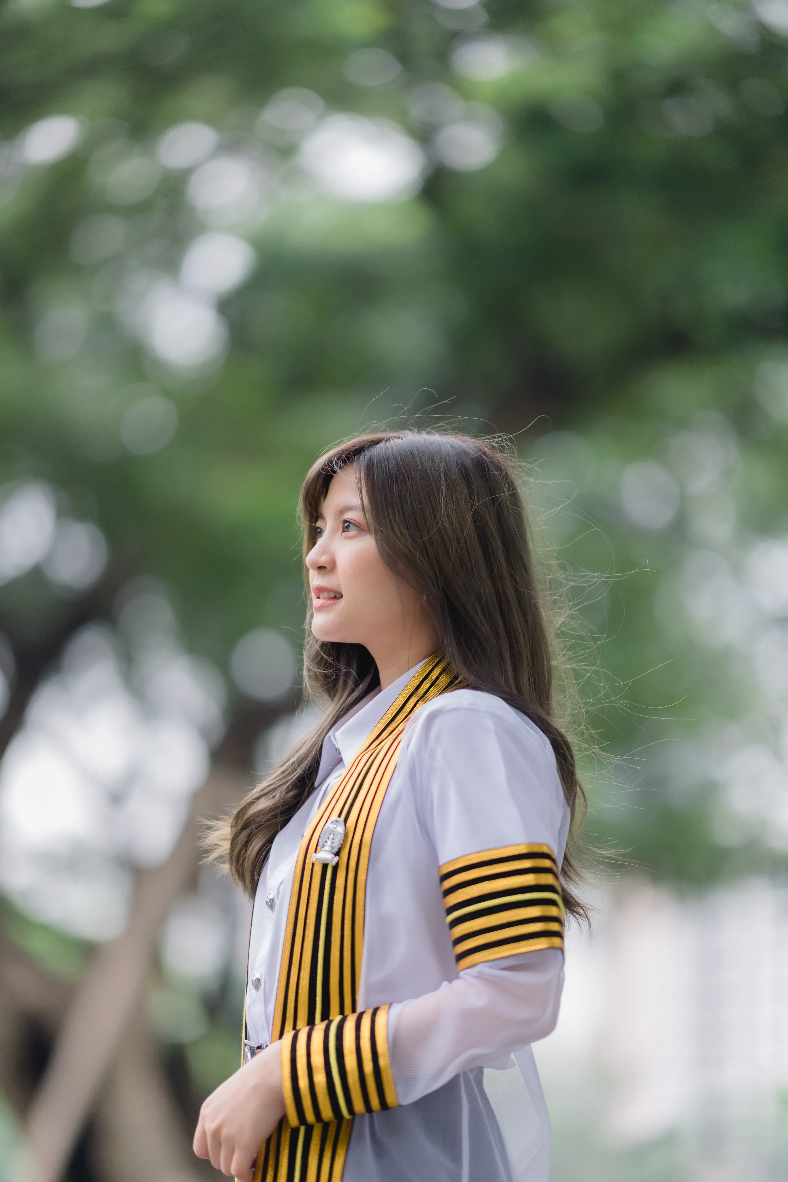A graduate in a striped academic gown gazing to the side in front of green trees