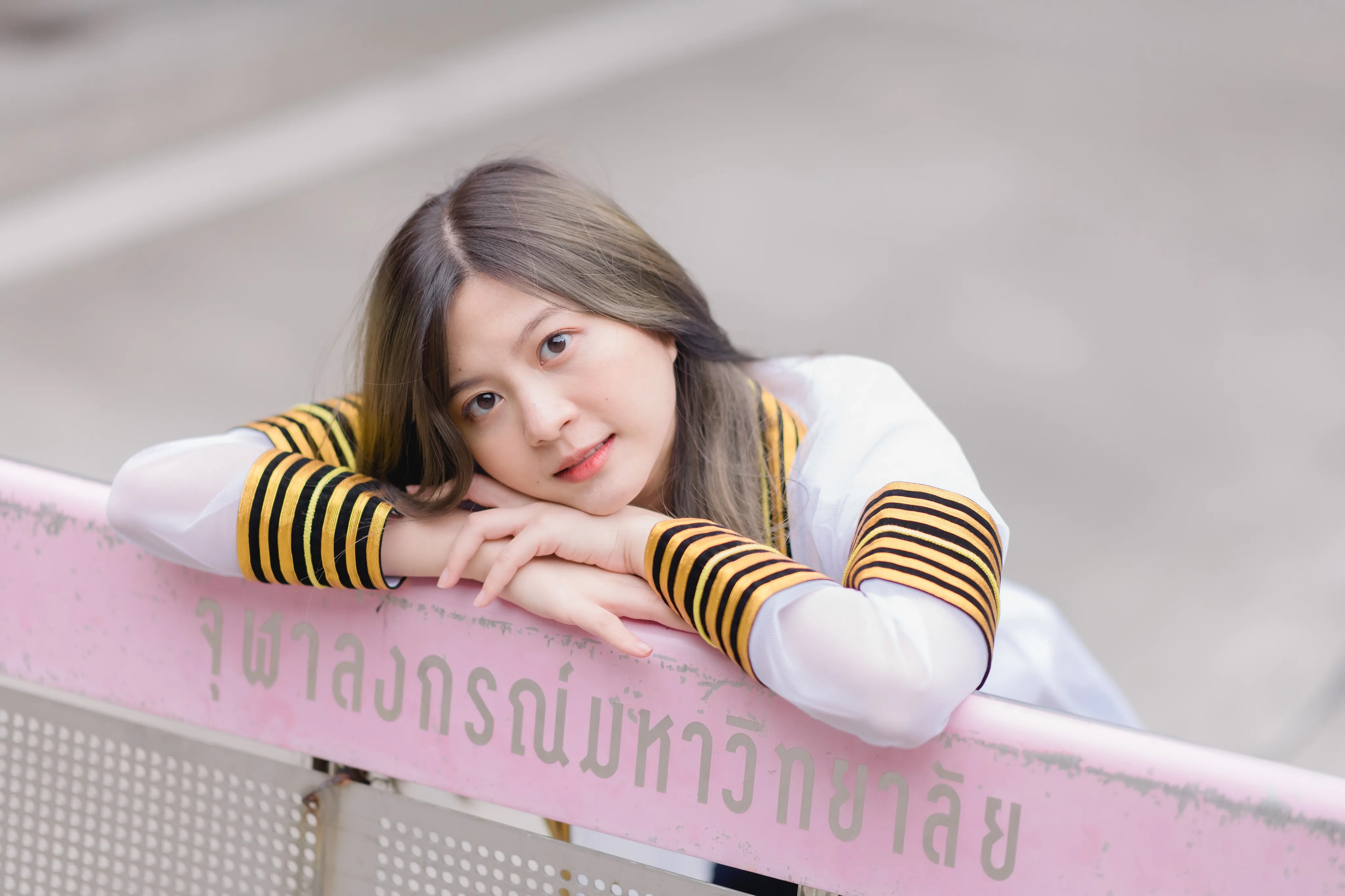 A graduate in a striped academic gown resting her head on her folded arms on a pink railing