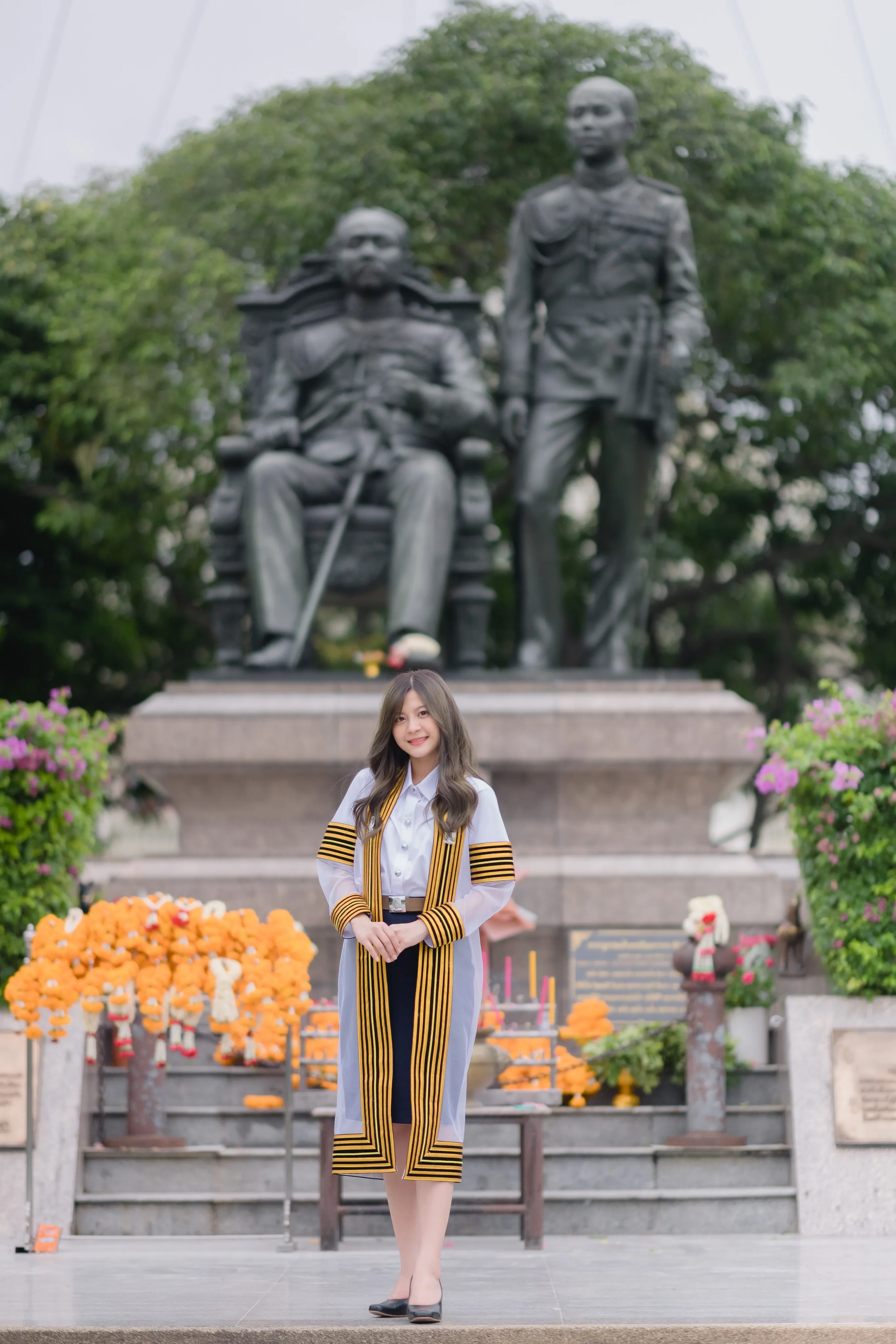 A graduate in a striped academic gown standing before a monument of two bronze statues with orange flower offerings
