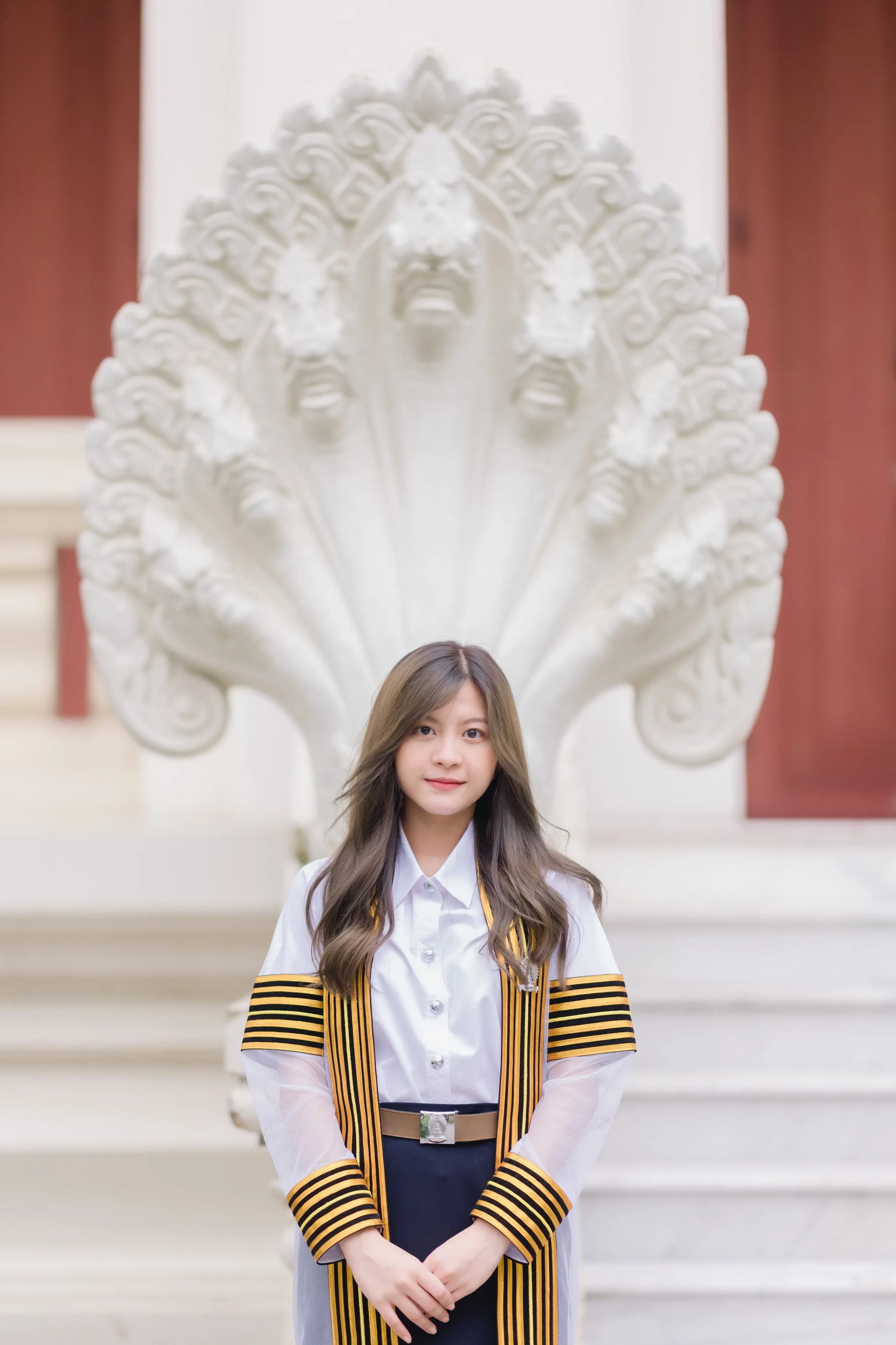 A graduate in a striped academic gown standing before a white carved serpent sculpture