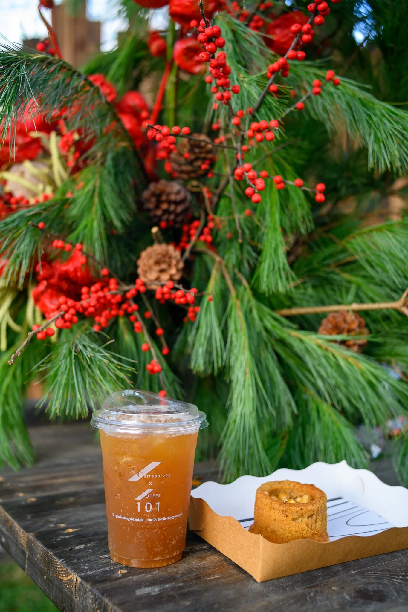 An iced drink and a small cake on a wooden table in front of a pine-and-berry arrangement