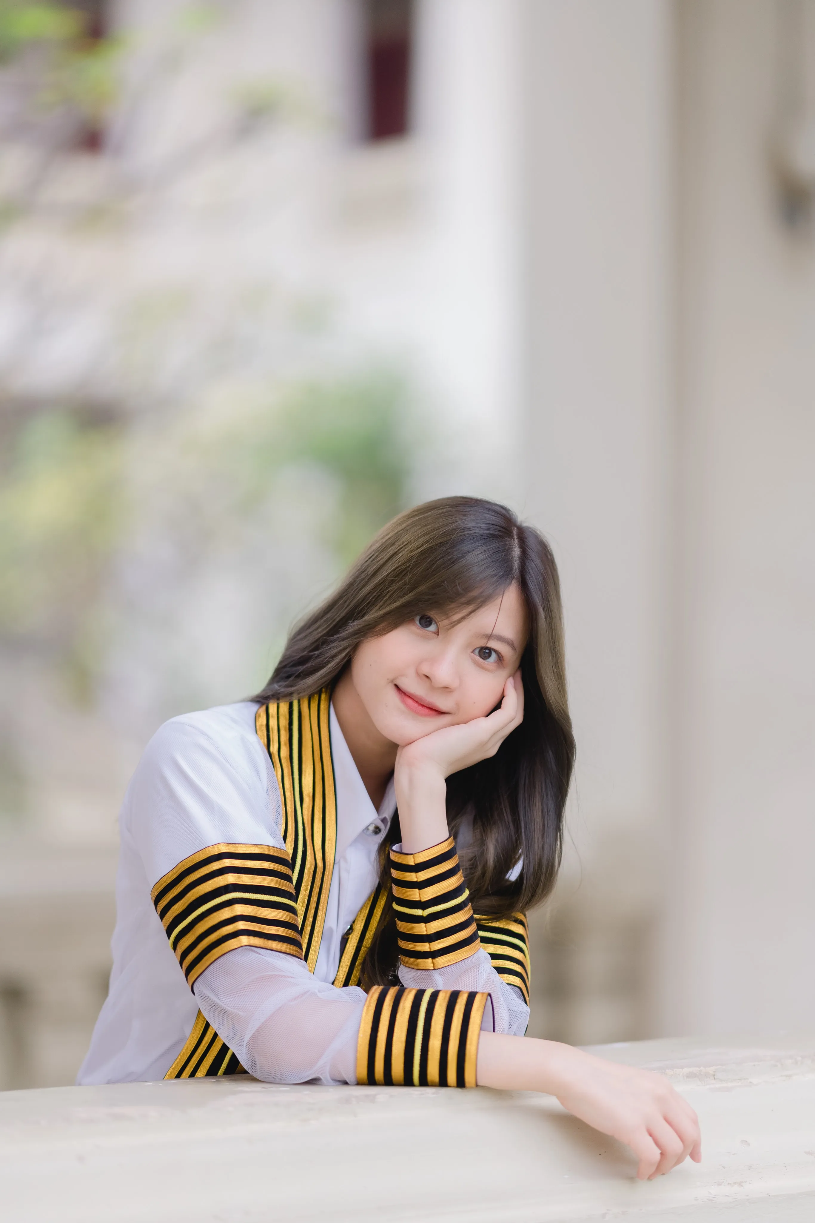 A smiling graduate in a striped academic gown resting her chin on her hand at a stone ledge