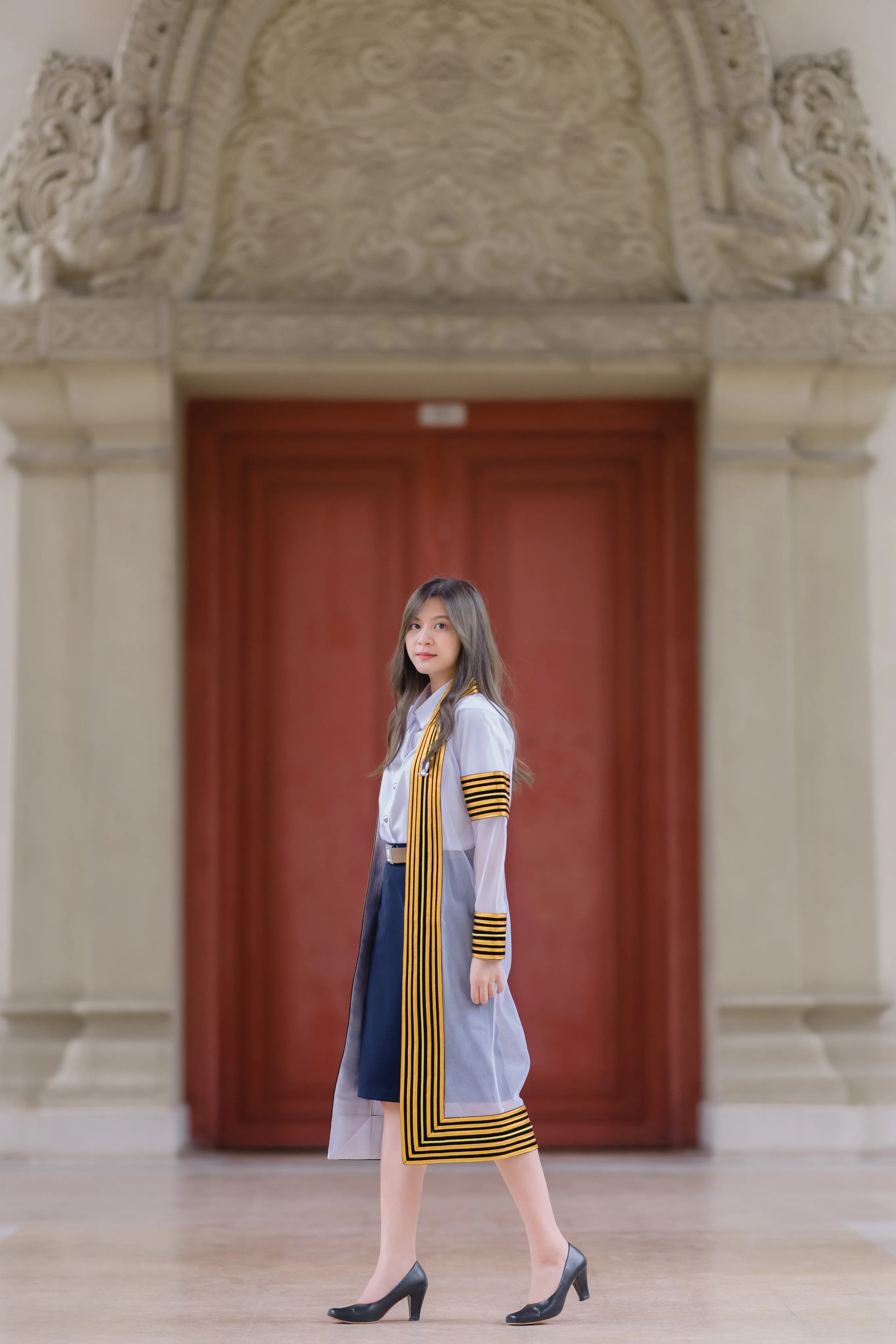 A graduate in a striped academic gown walking past a tall red door in a carved stone doorway