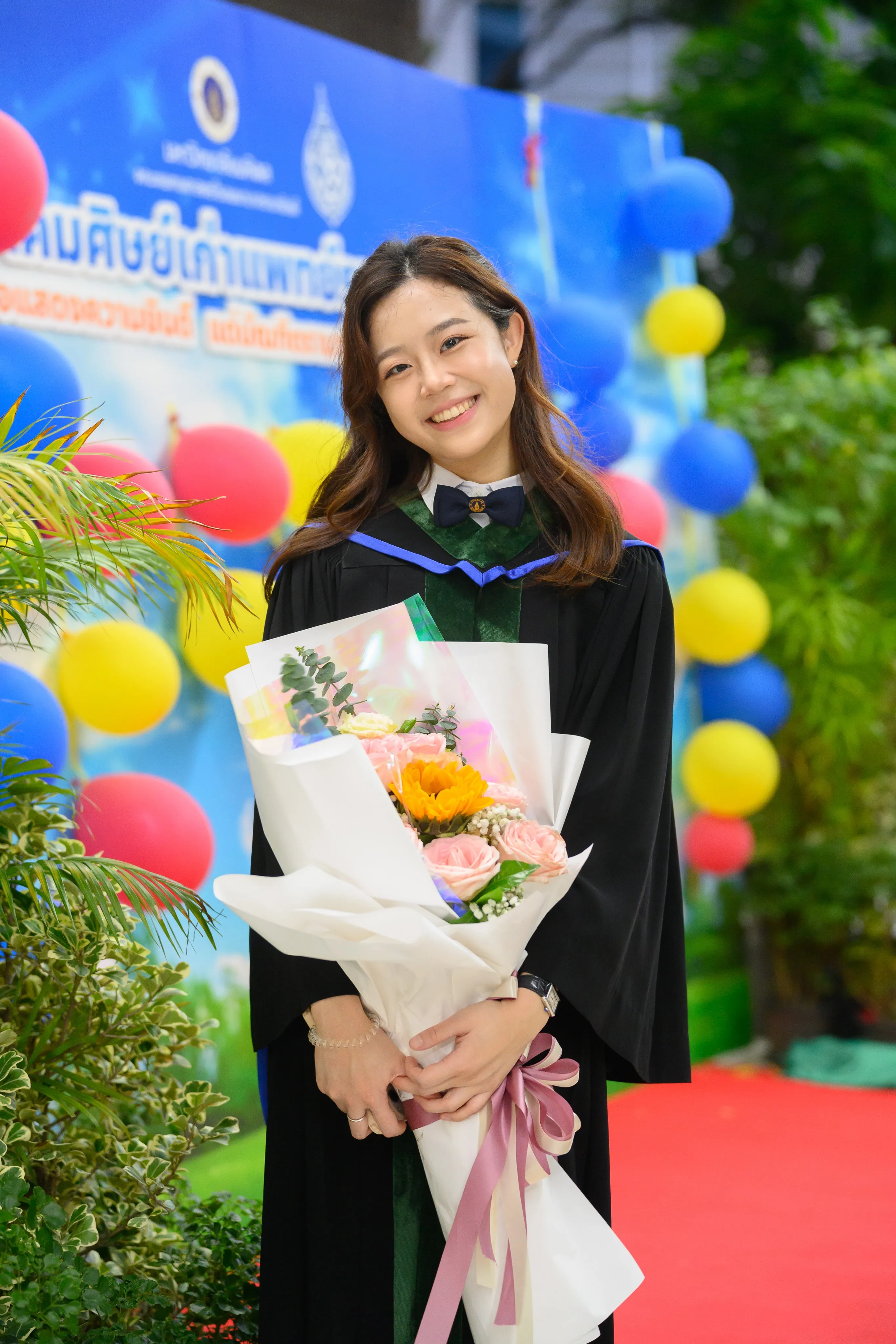 A smiling graduate in a black gown holding a large bouquet in front of a balloon-decorated backdrop