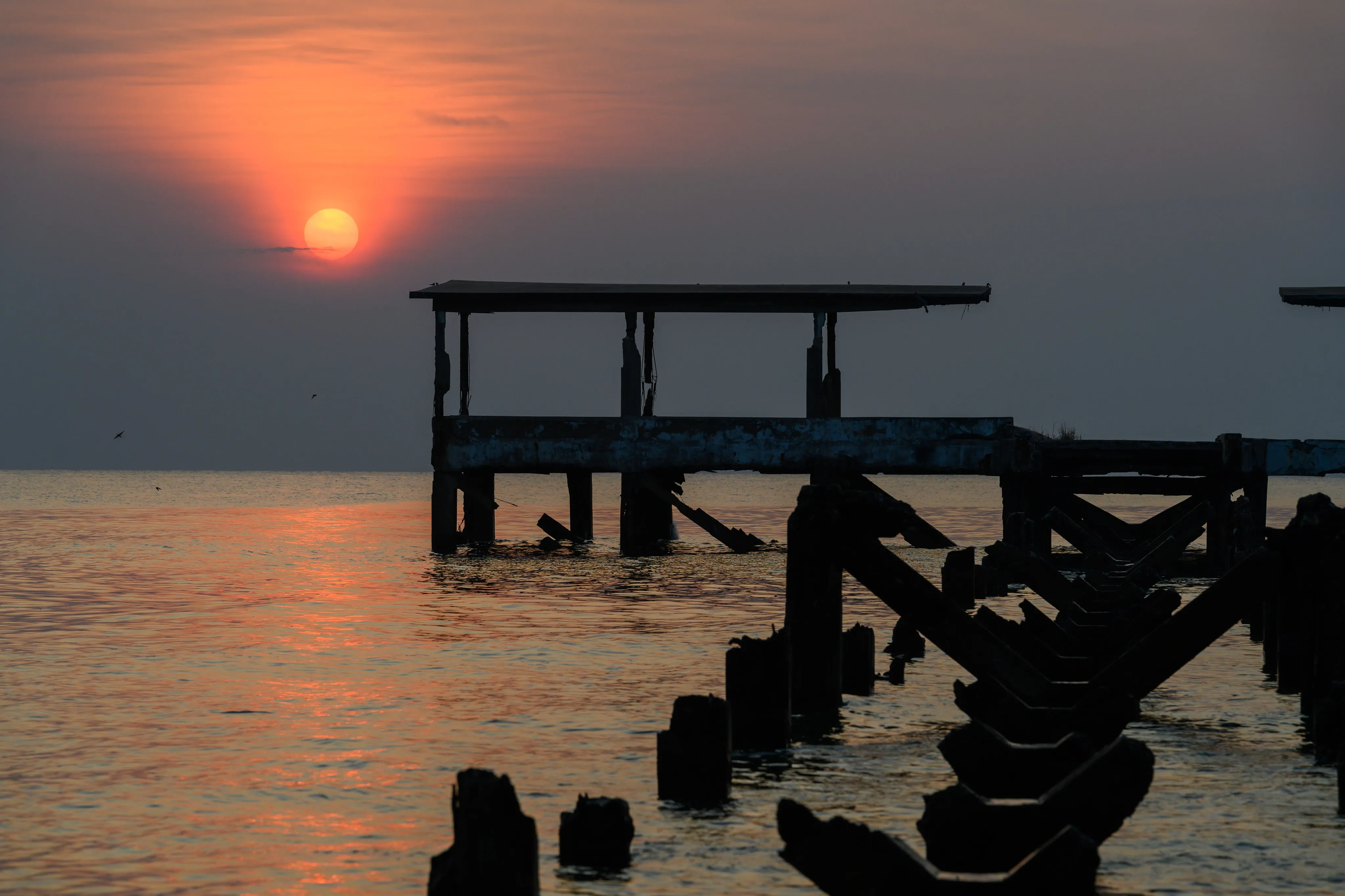 The sun setting in a red sky over the sea behind the ruins of an old wooden pier