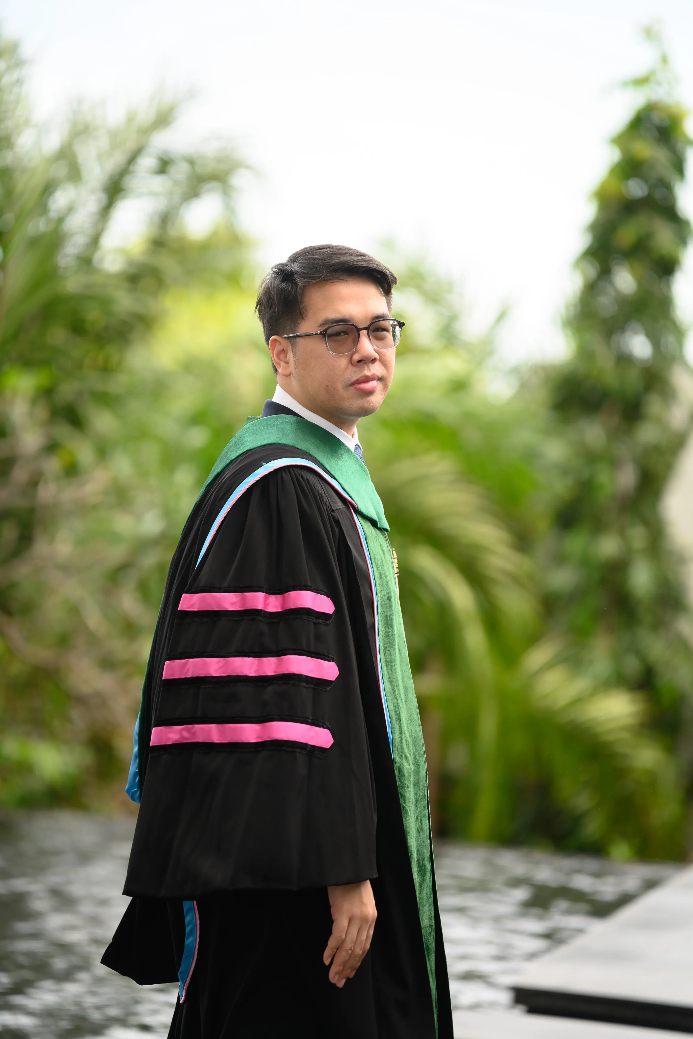 A man in glasses wearing a black doctoral gown with pink velvet bars in a garden