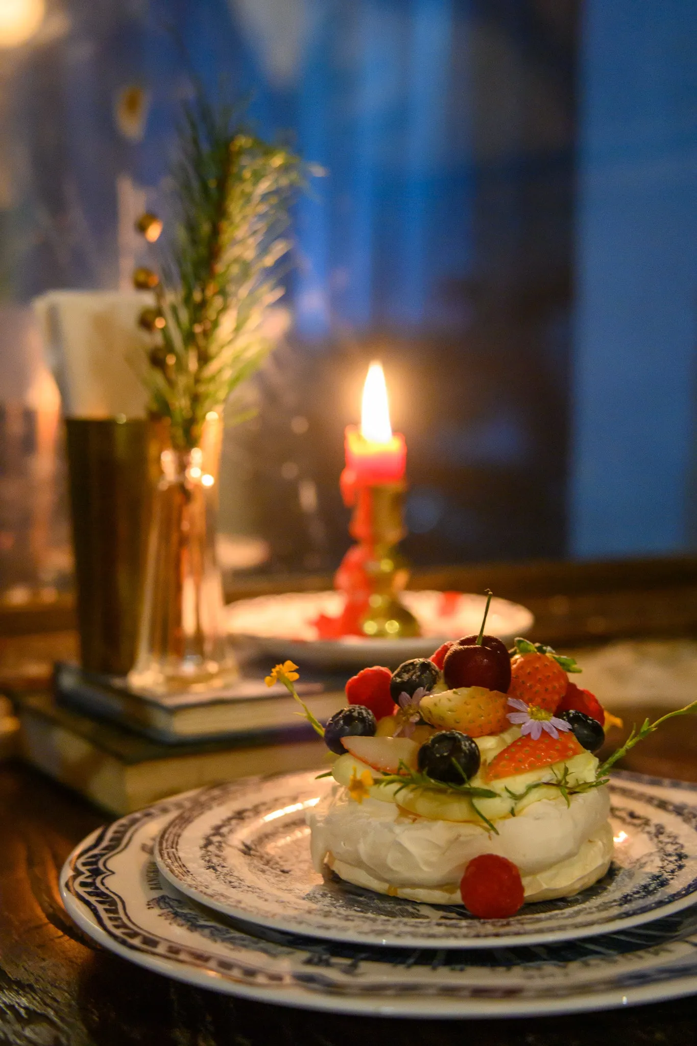 A fruit-topped pavlova on patterned plates beside a lit red candle