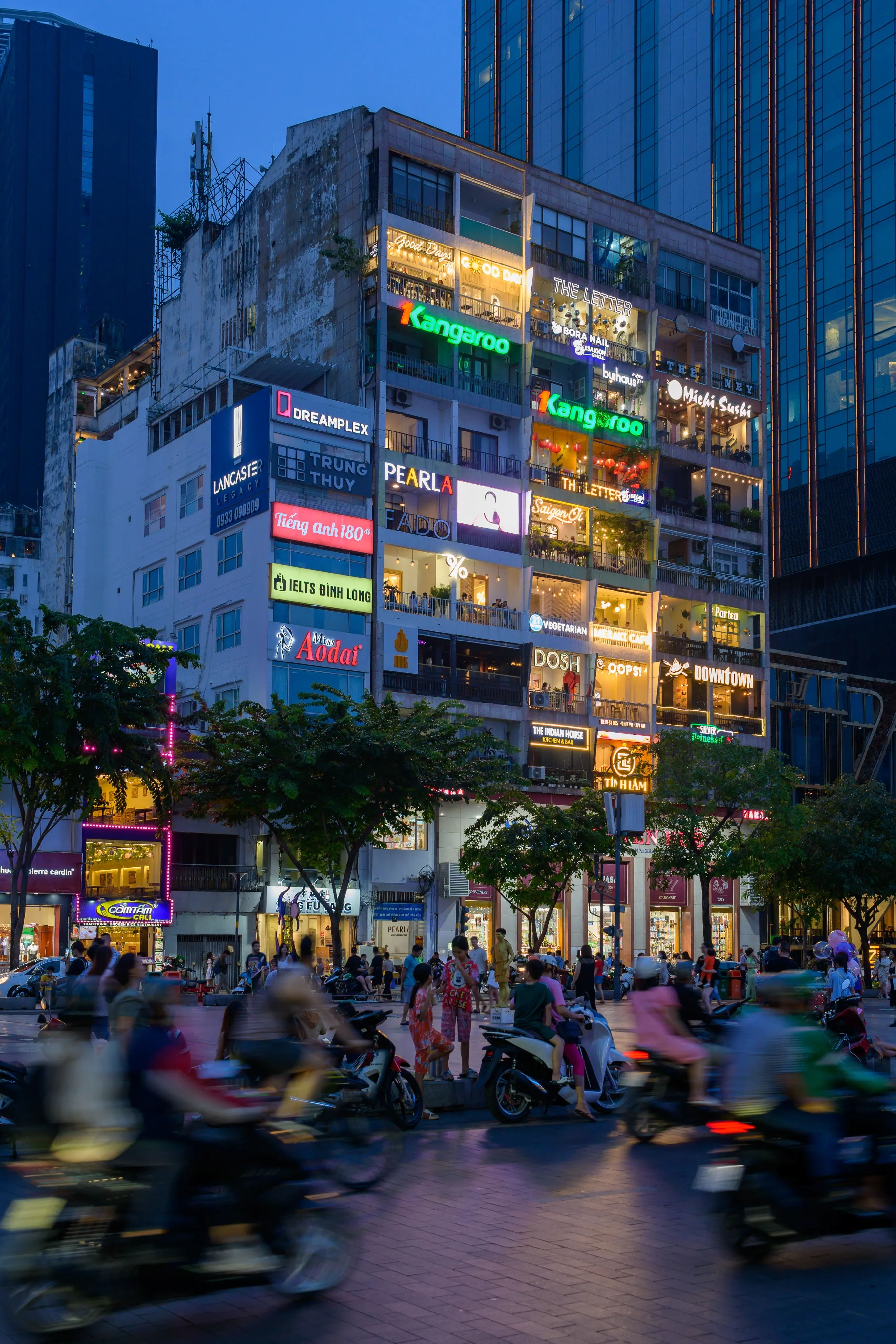 A neon-lit apartment block of shops and cafés at dusk, with motorbikes streaming past in the street below