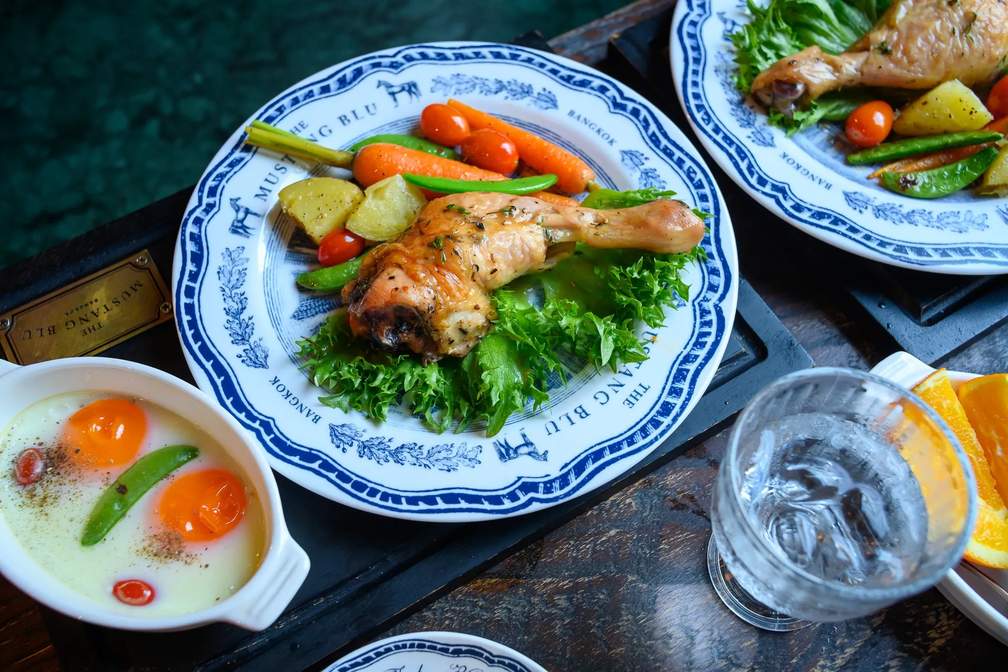Roast chicken with vegetables on a blue-and-white plate beside baked eggs and a glass of water