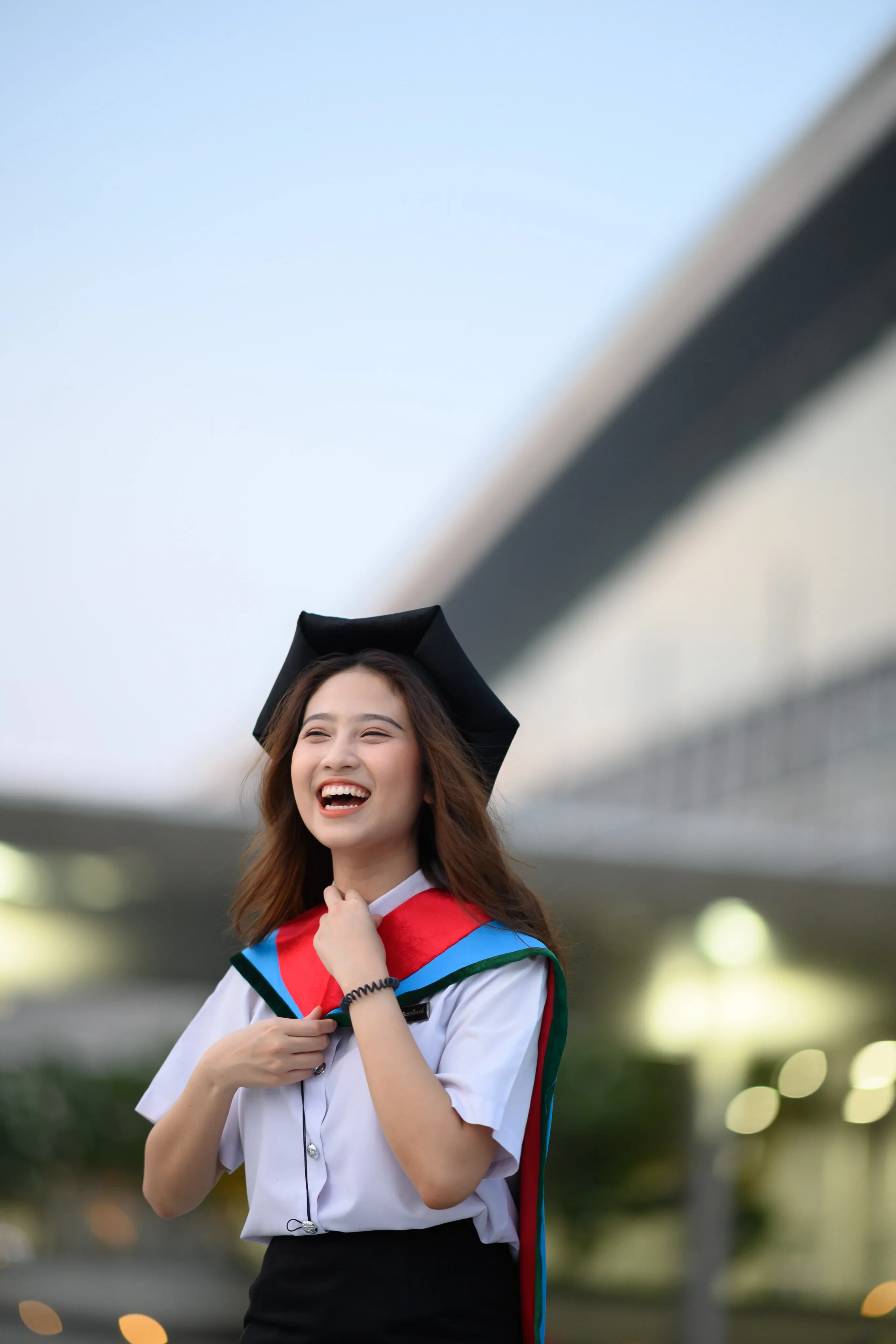 A laughing graduate in a mortarboard with a red-and-blue hood in front of a blurred building