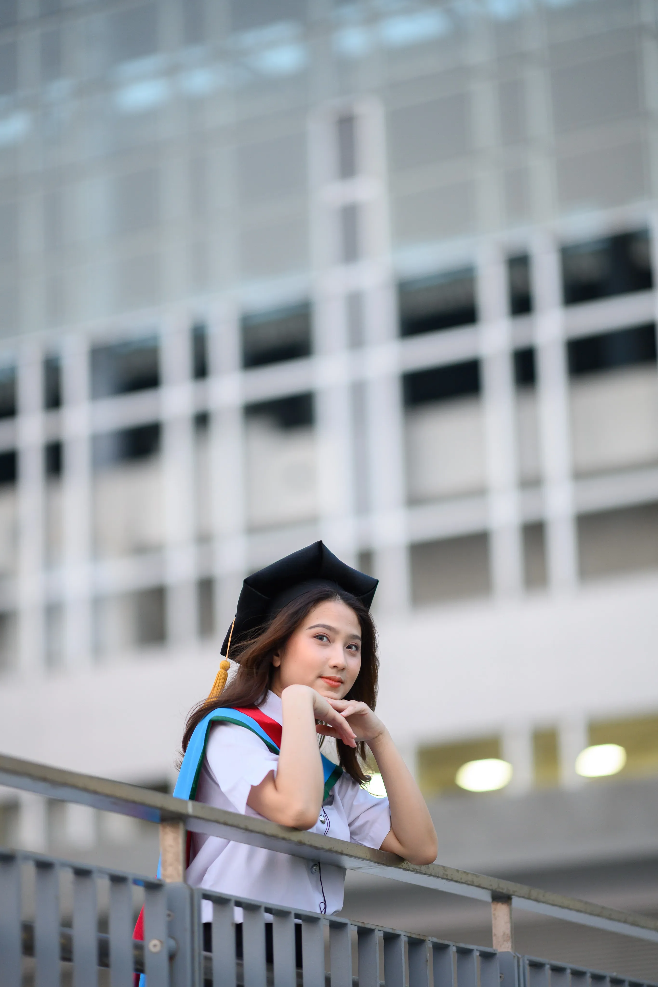 A graduate in a mortarboard resting her chin on her hands at a railing outside a glass building