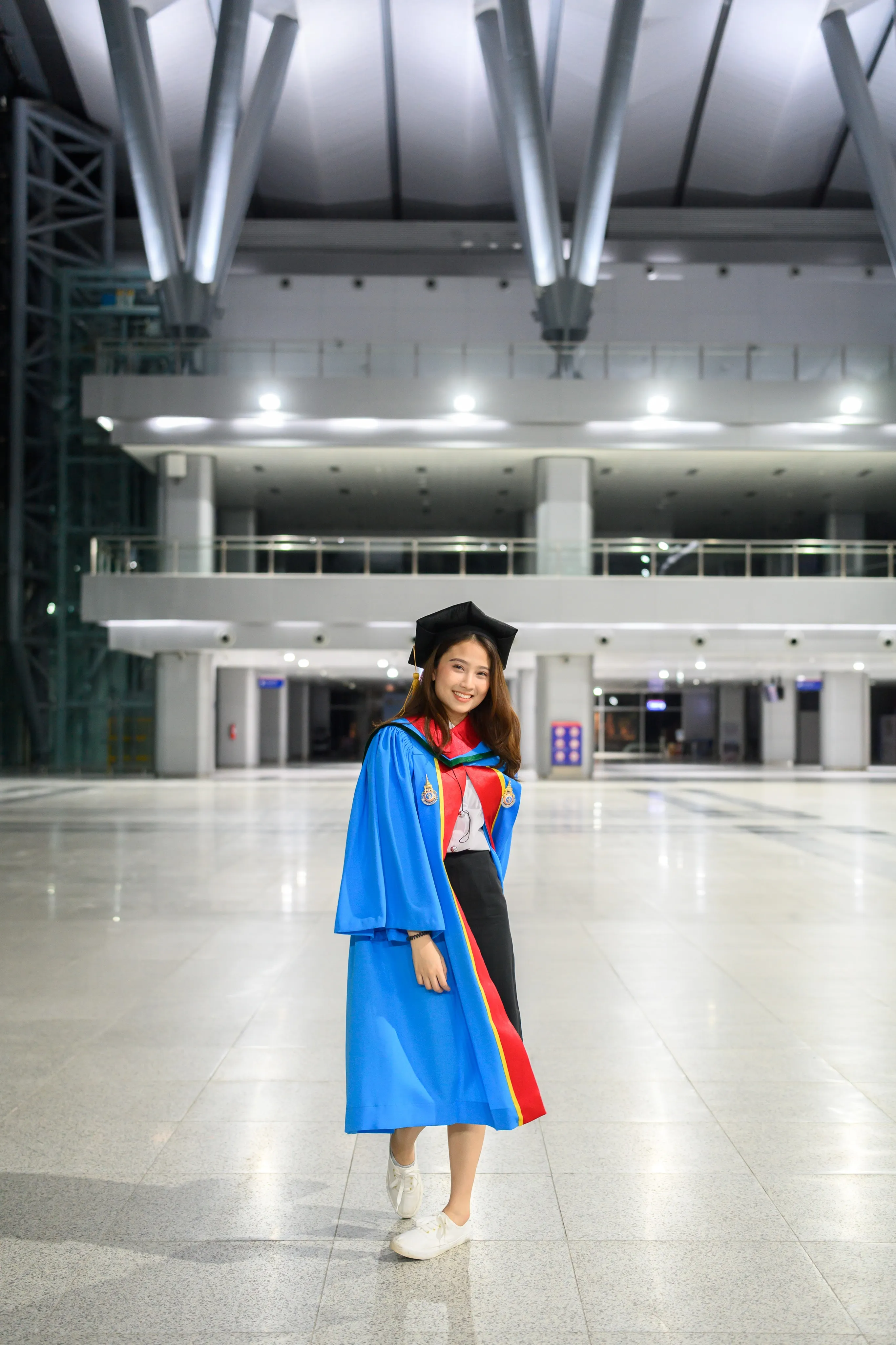 A graduate in a mortarboard and blue gown standing in a large, brightly lit hall