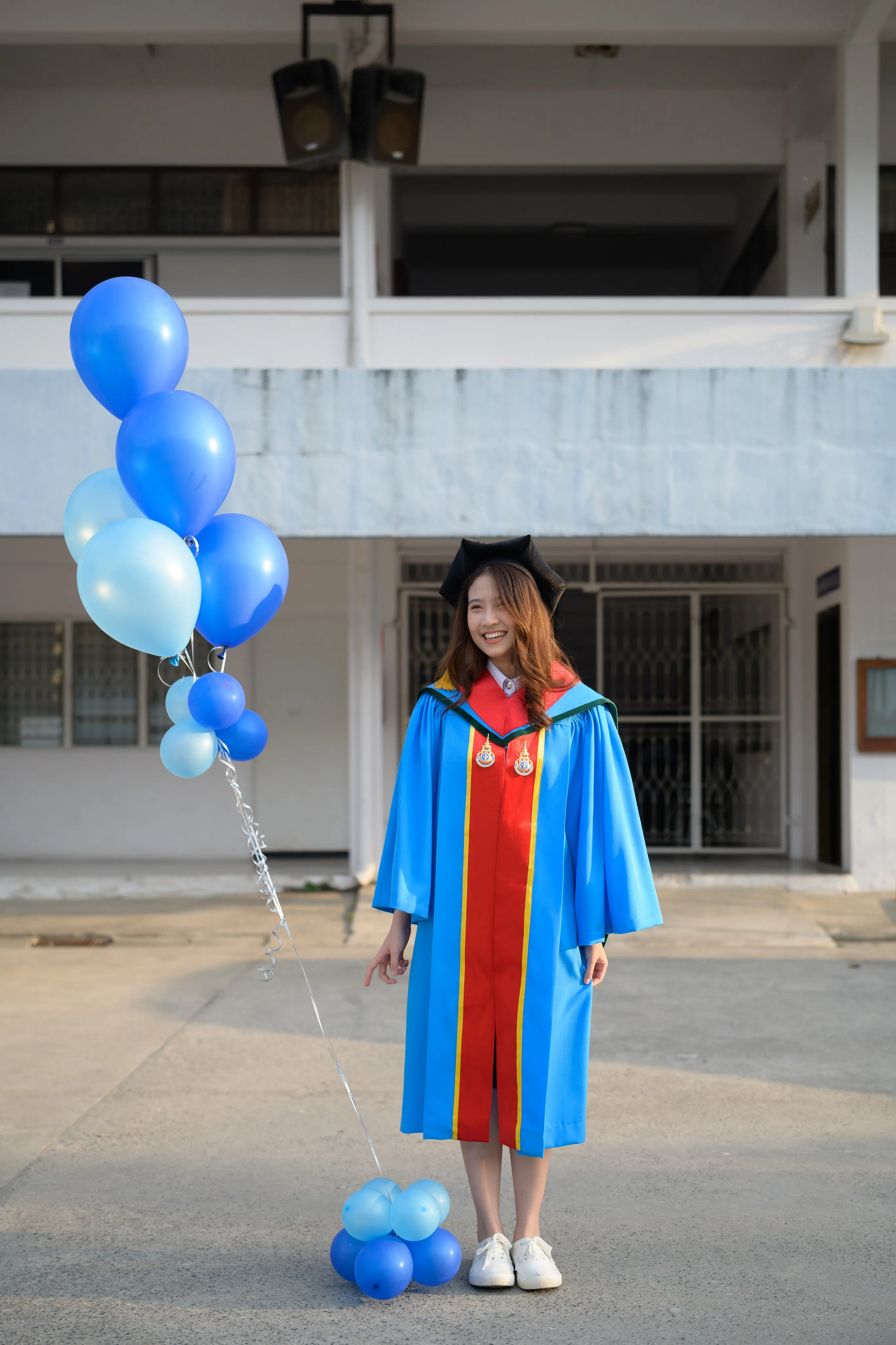A graduate in a mortarboard and blue gown standing with a bunch of blue balloons outside a building