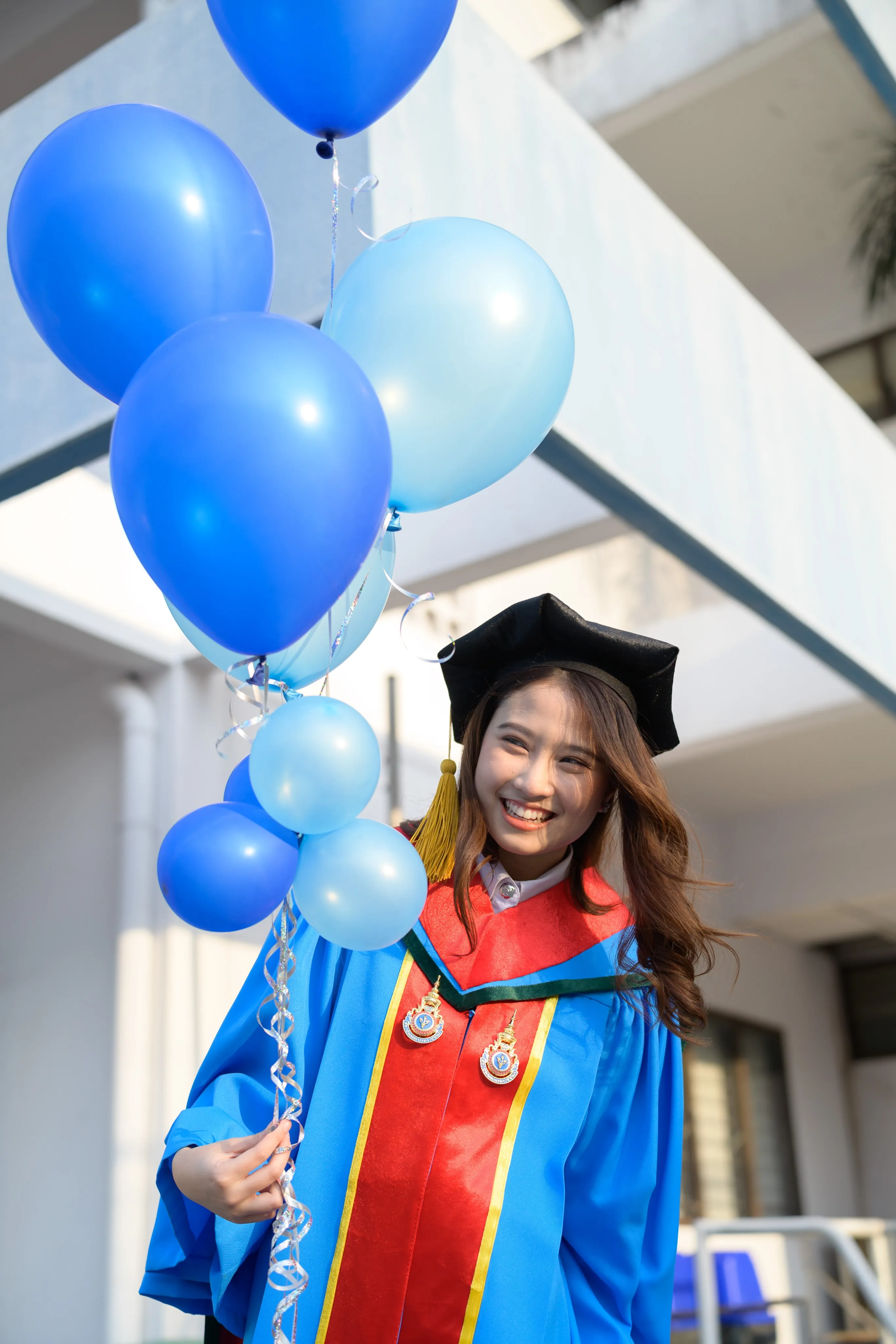 A smiling graduate in a mortarboard and blue gown holding a bunch of blue balloons