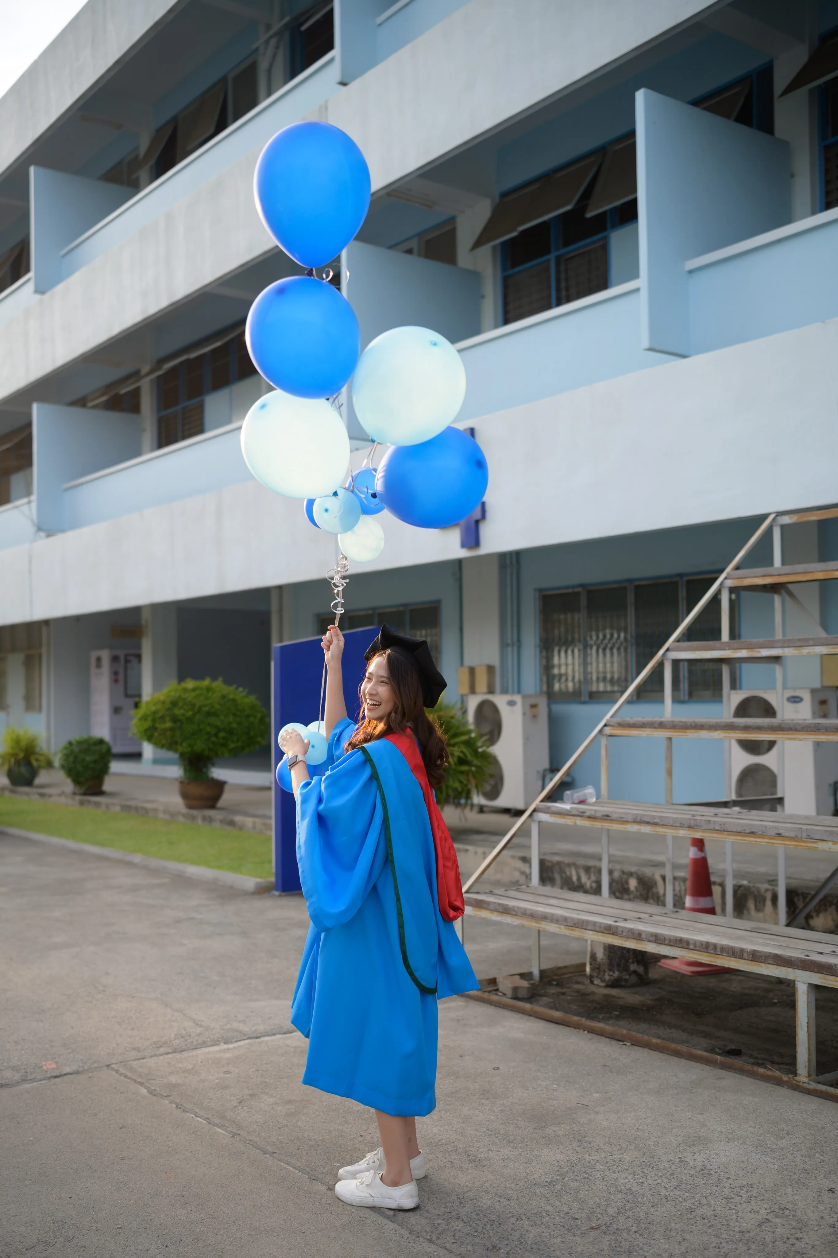 A graduate in a blue gown holding up blue balloons in a courtyard beside a pale blue building