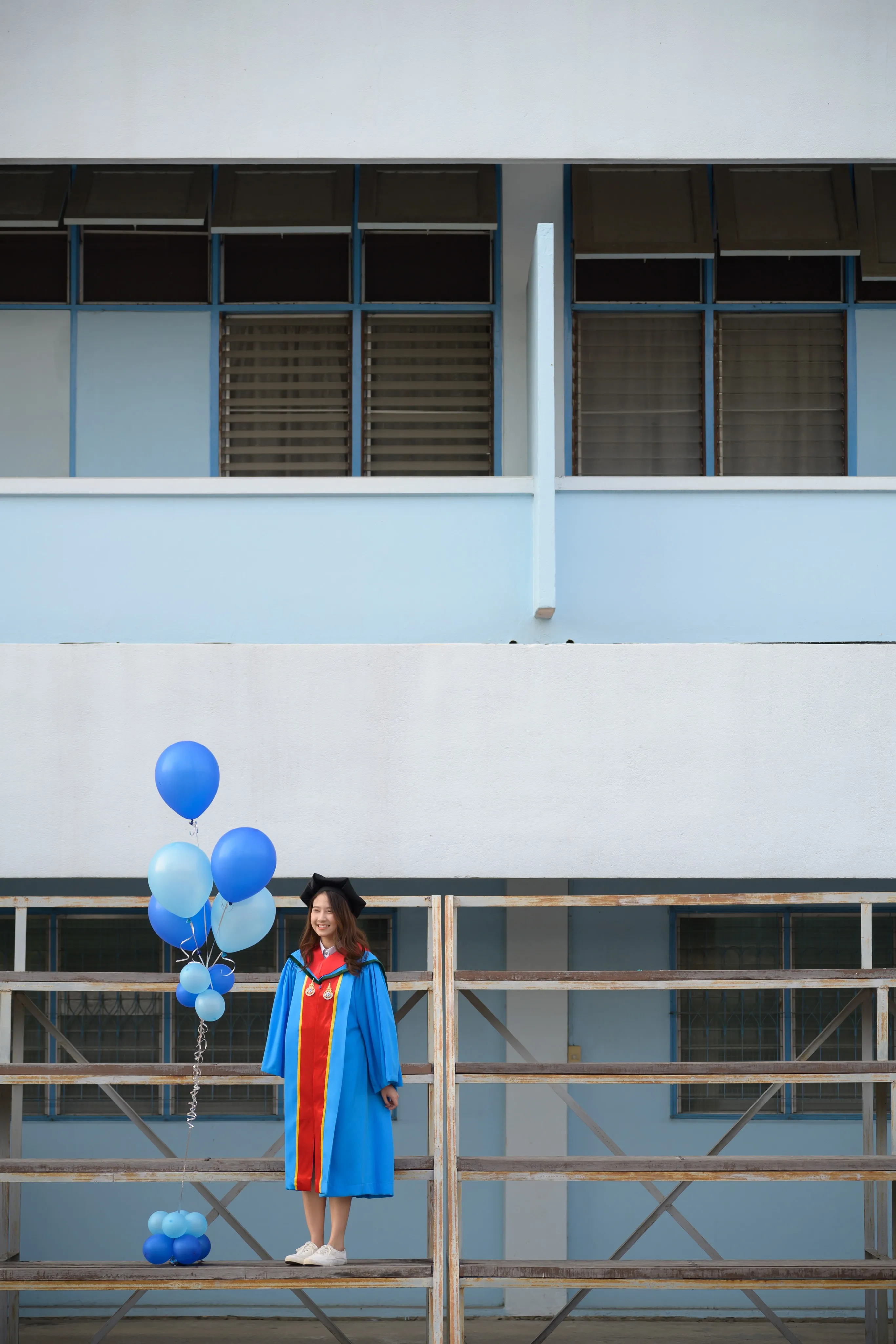 A graduate in a blue gown standing on wooden bleachers with blue balloons against a pale blue building