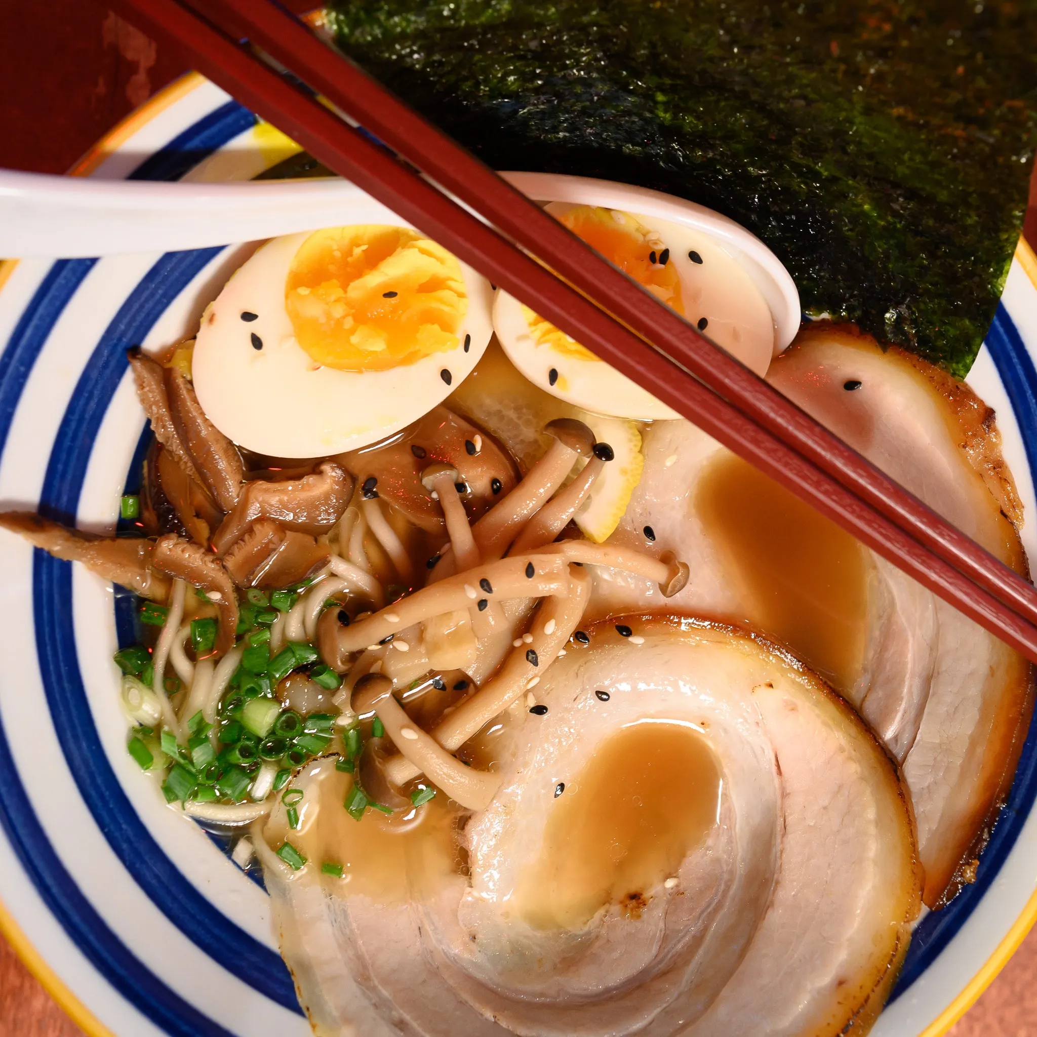 A bowl of ramen with chashu pork, soft-boiled eggs, mushrooms, and nori, with chopsticks