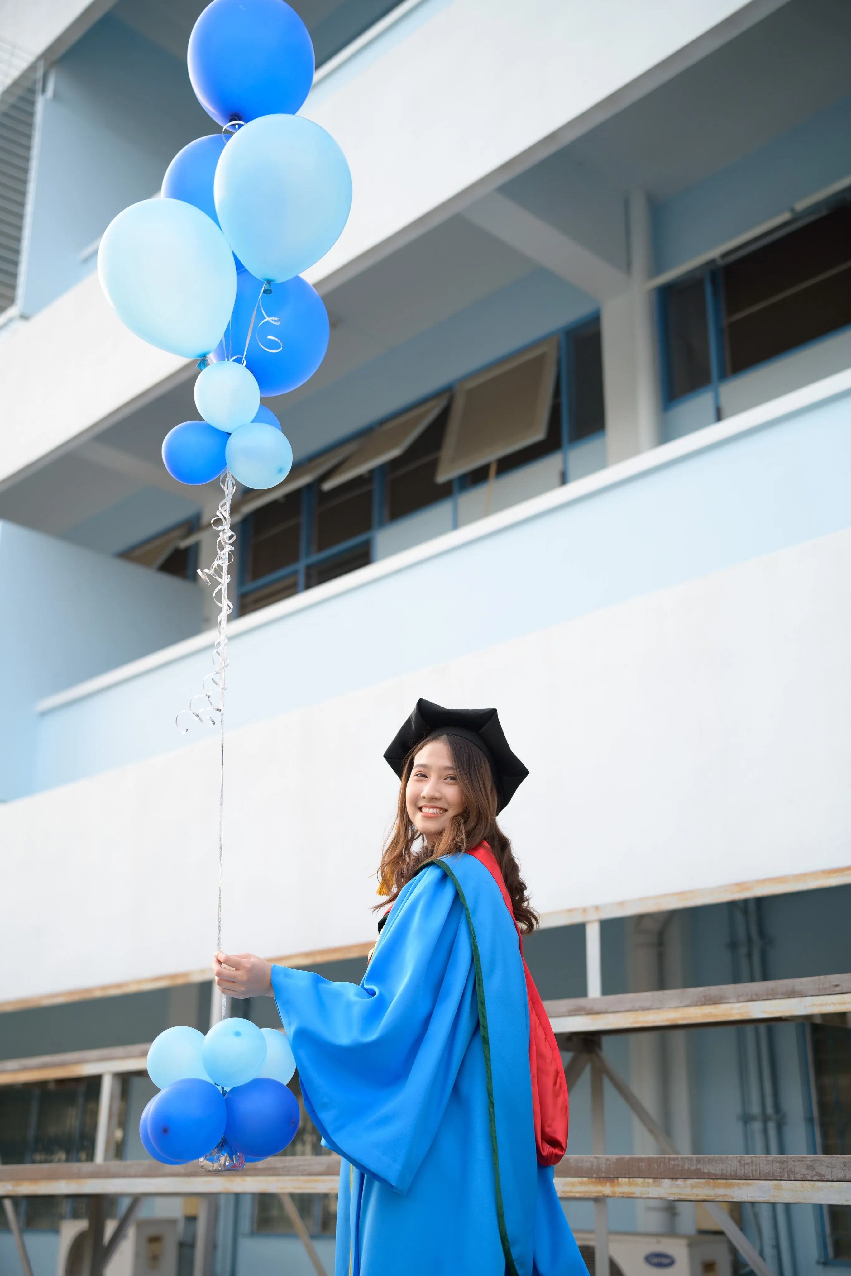 A smiling graduate in a mortarboard and blue gown holding blue balloons on wooden bleachers