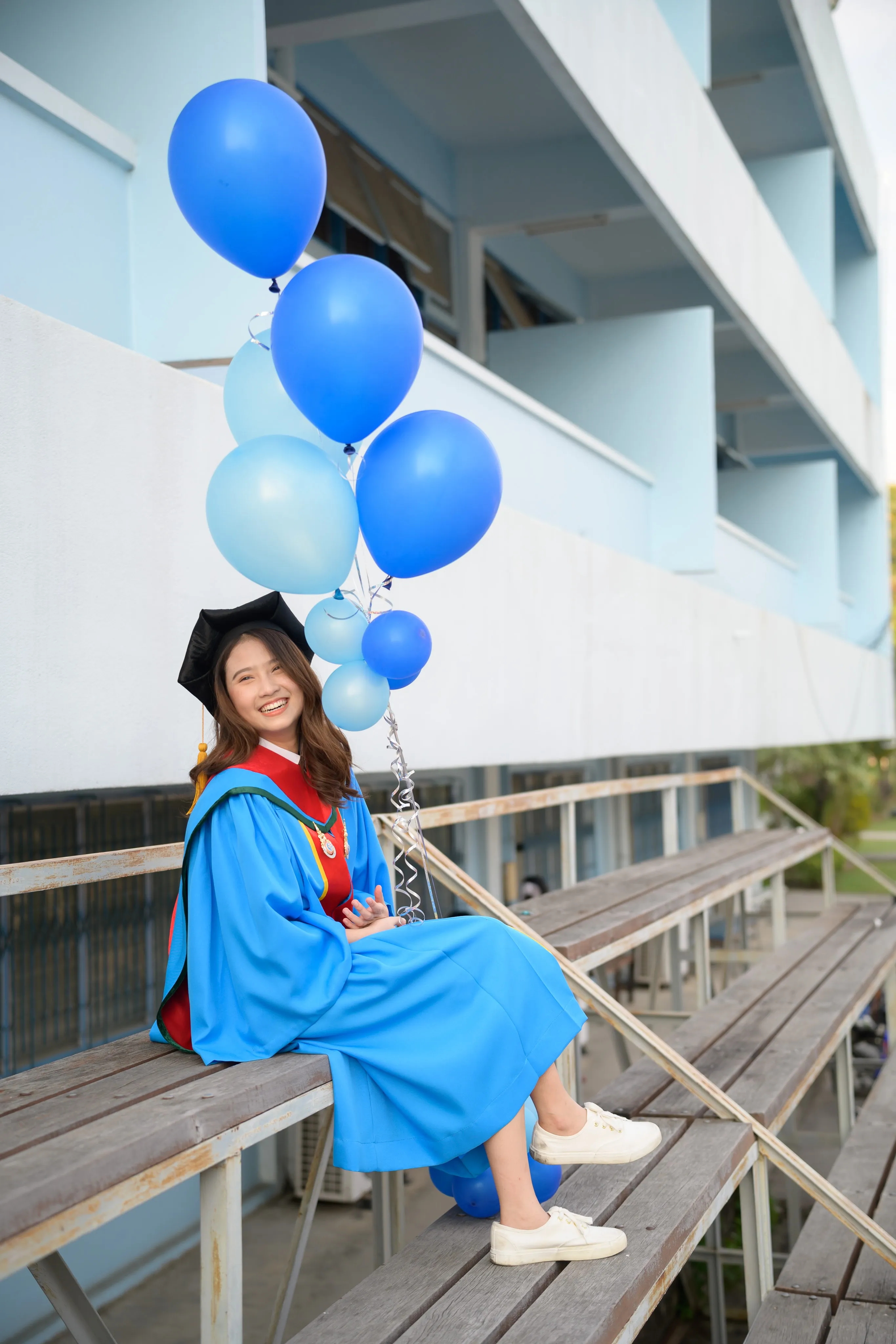 A smiling graduate in a blue gown sitting on wooden bleachers with blue balloons