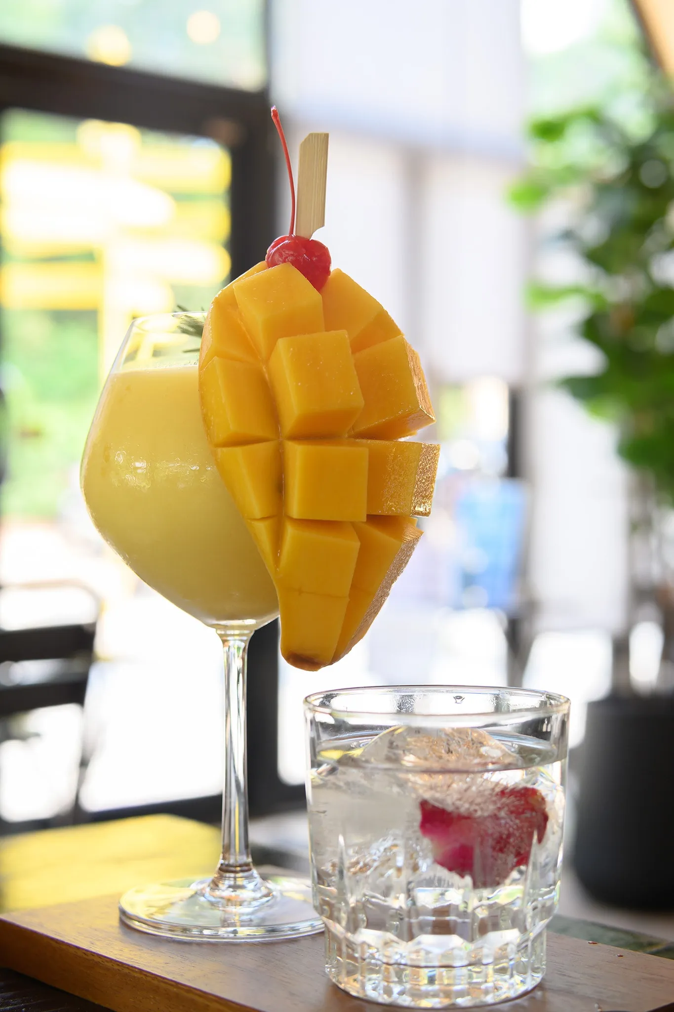 A mango smoothie garnished with a carved mango and a cherry beside a glass of iced water