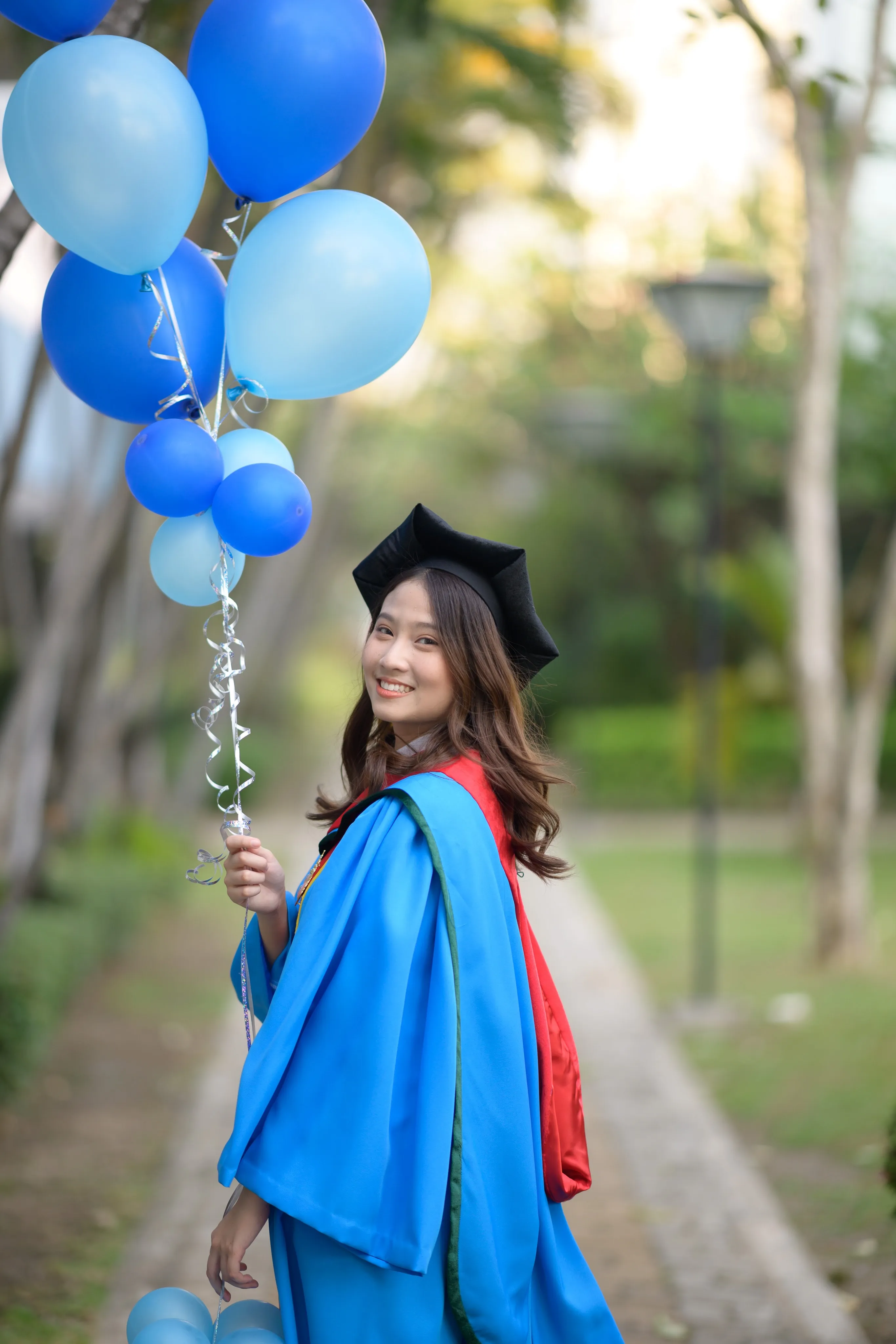 A smiling graduate in a mortarboard and blue gown holding blue balloons on a garden path