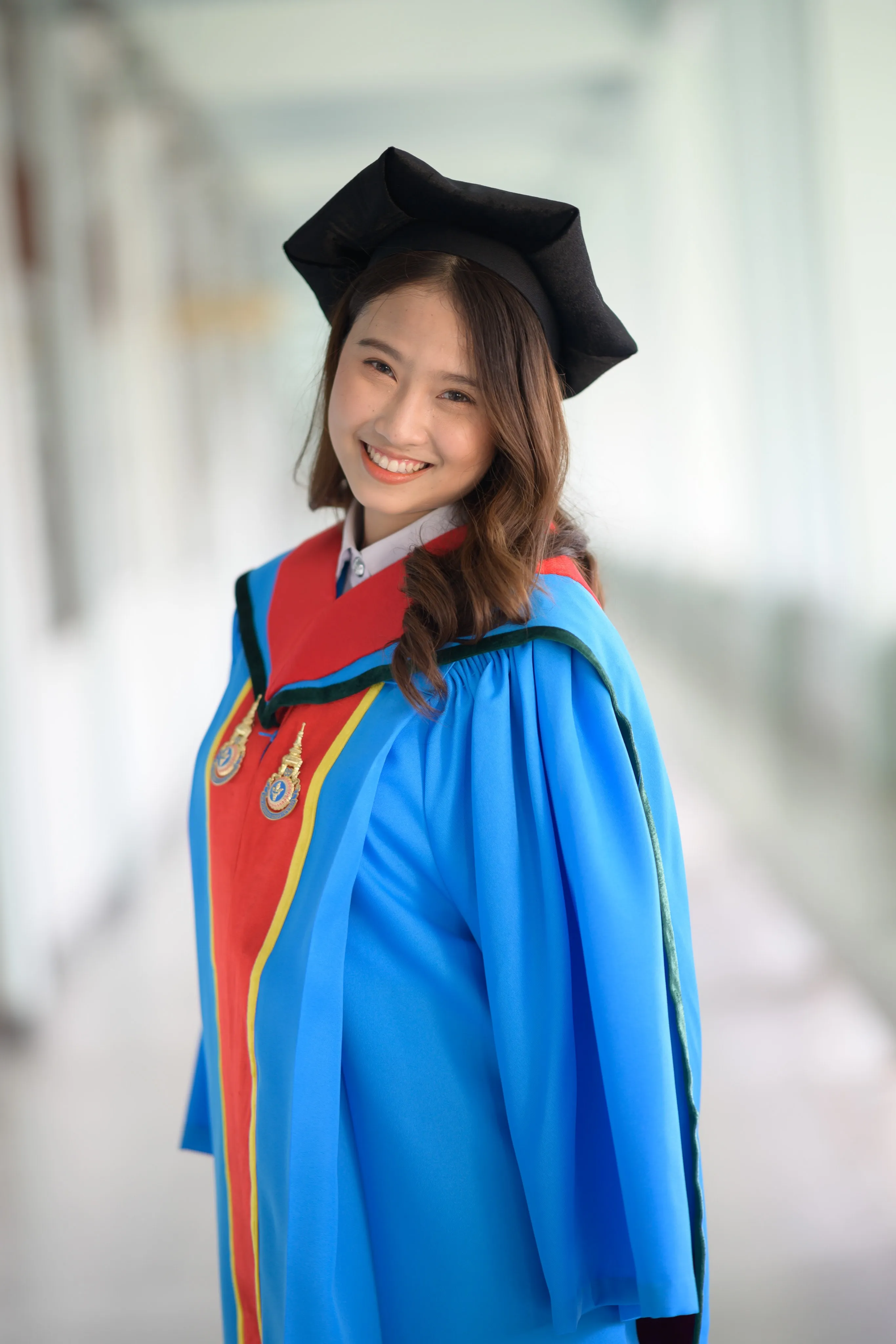A smiling graduate in a mortarboard and blue-and-red gown in a bright corridor