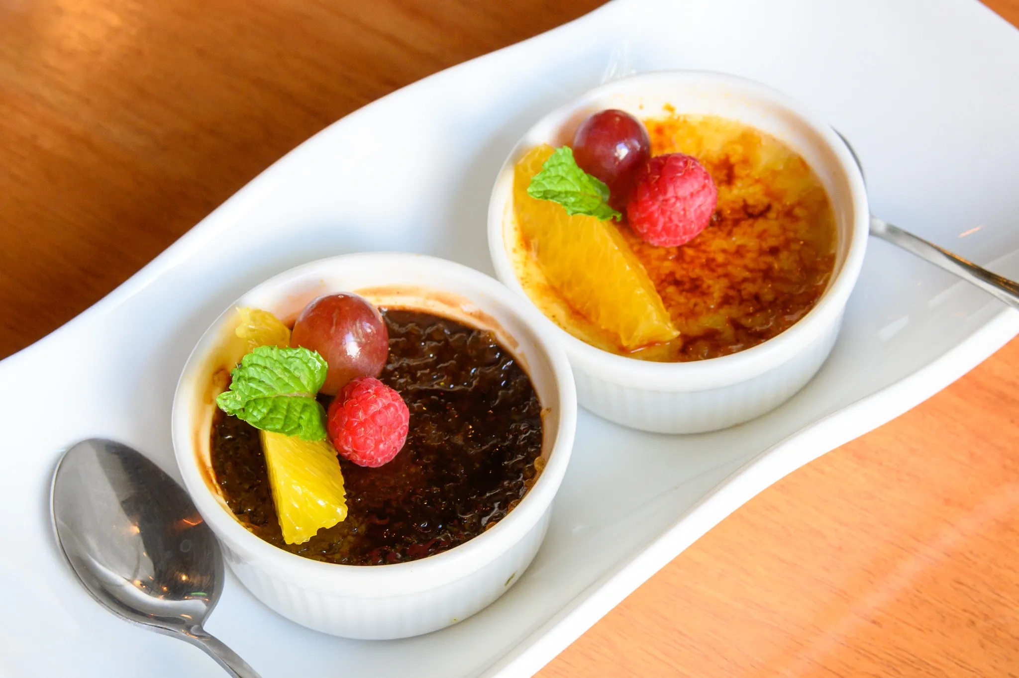 Two crème brûlées topped with fruit and mint on a white serving dish