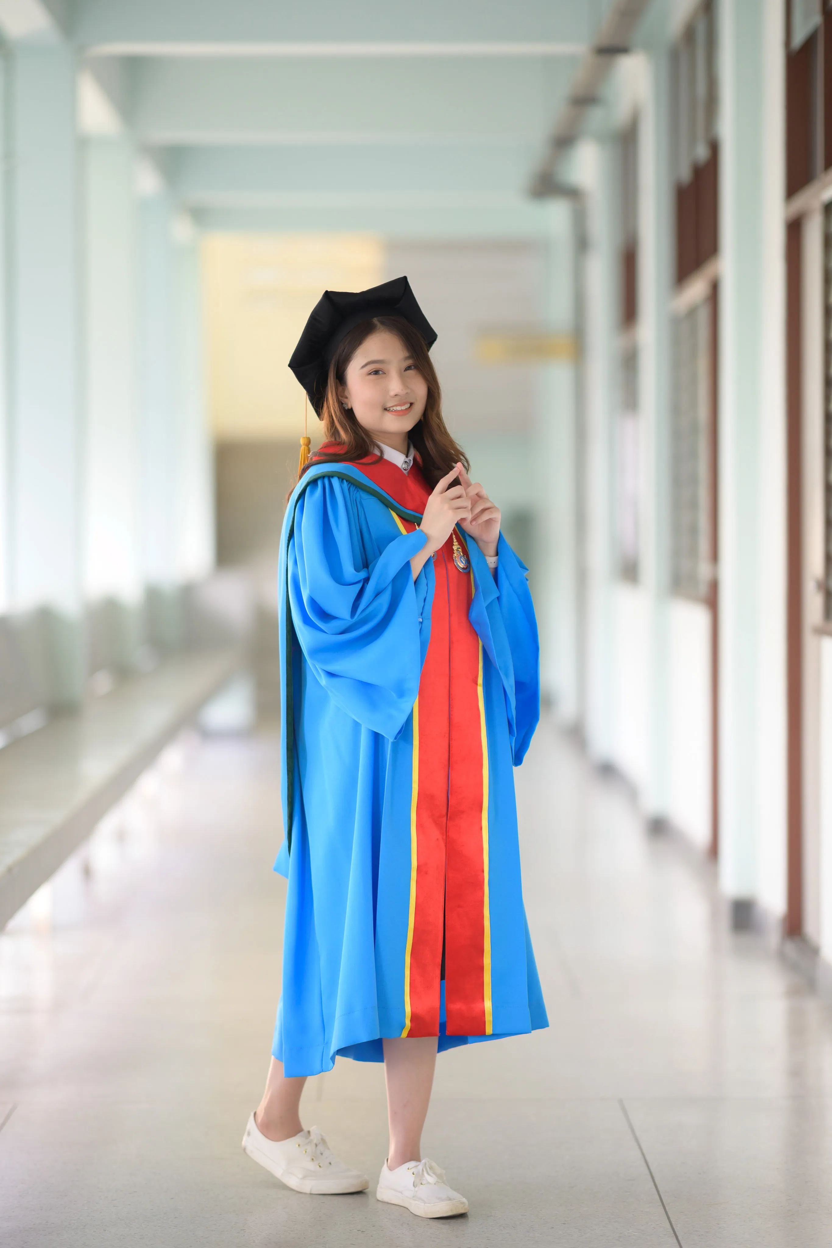 A graduate in a mortarboard and blue gown standing in a long open-air corridor