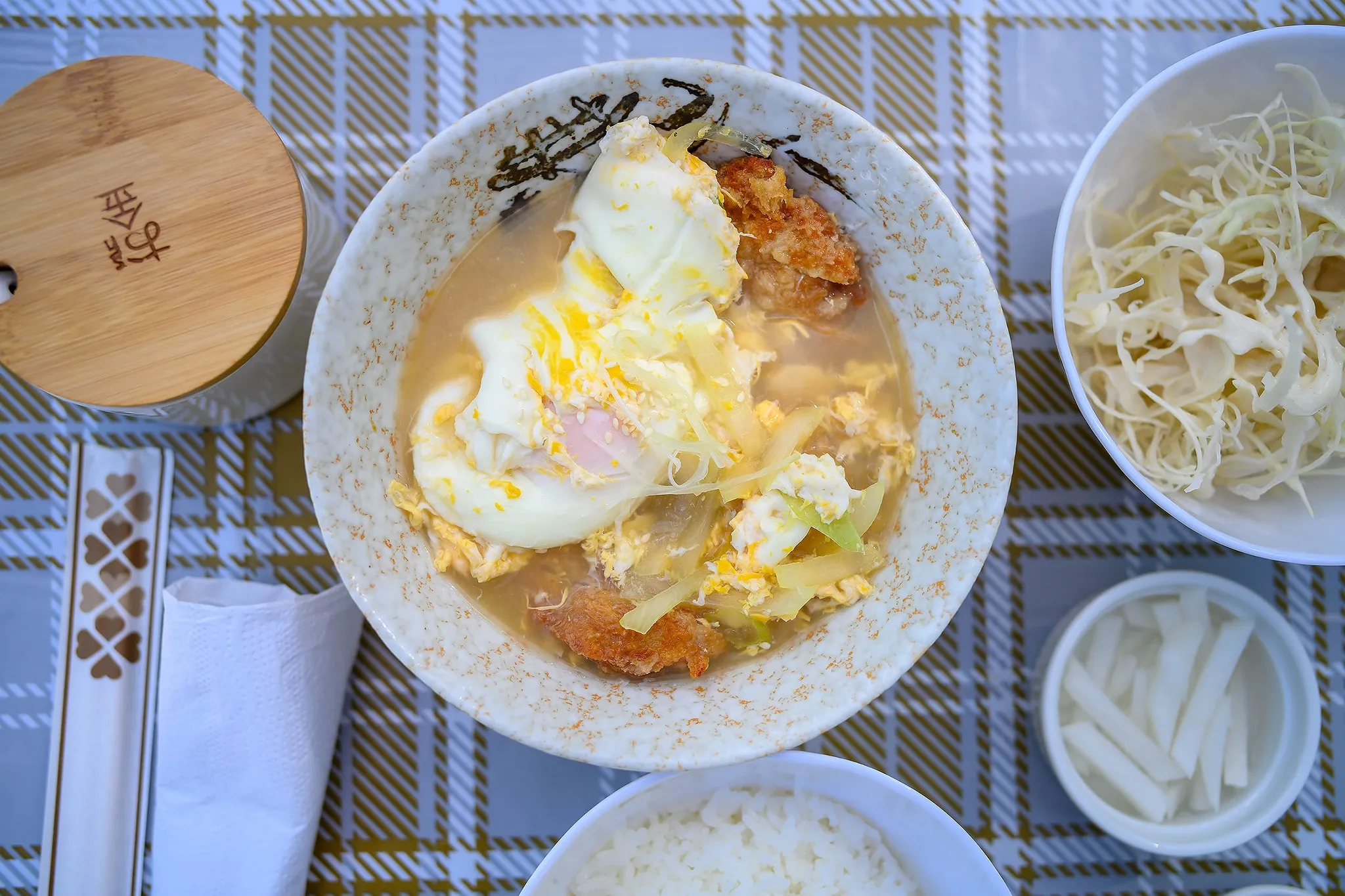 A bowl of fried pork cutlet with egg over broth beside side dishes of cabbage and pickled radish
