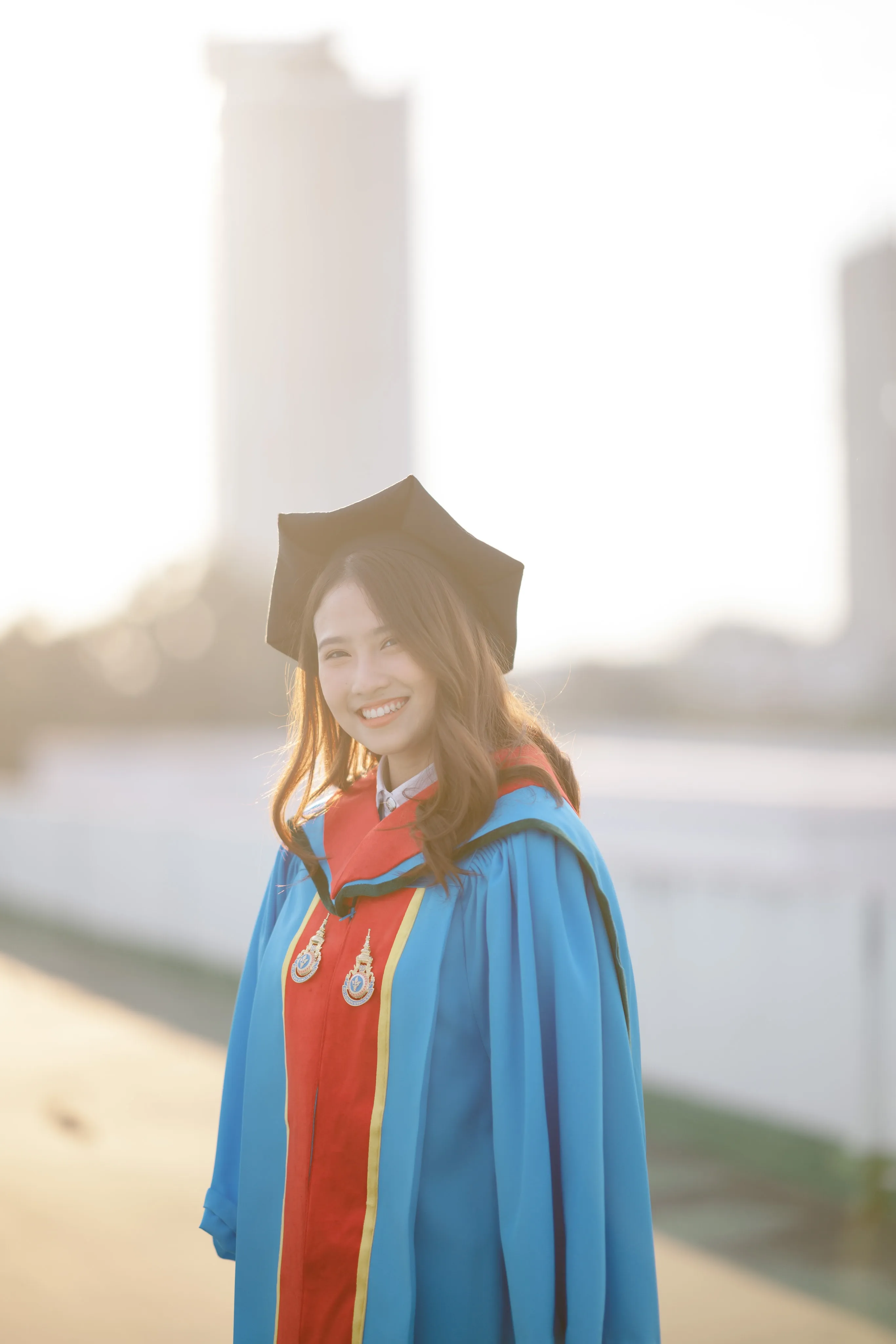 A smiling graduate in a mortarboard and blue gown on a rooftop in hazy sunlight