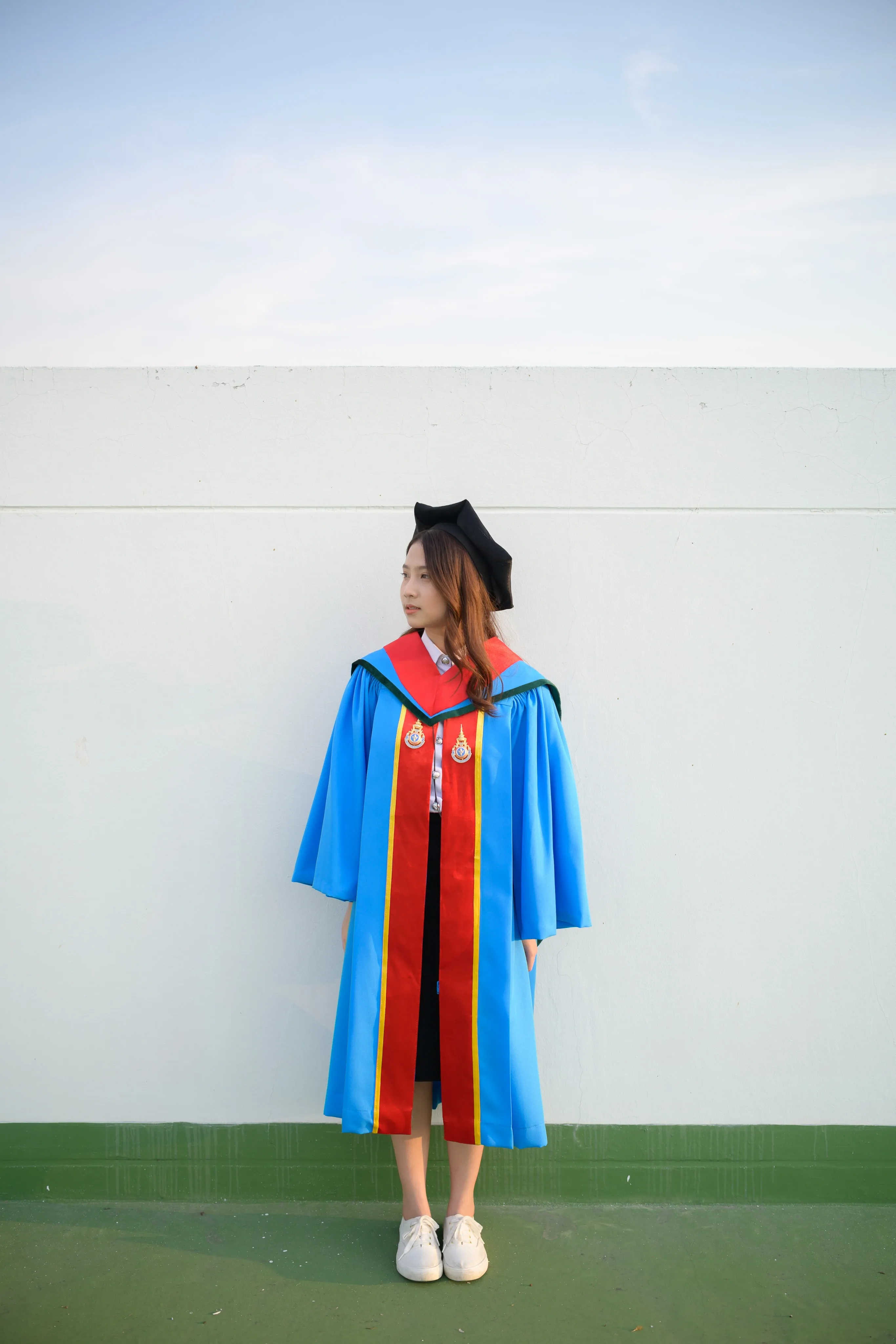 A graduate in a mortarboard and blue gown standing against a white rooftop wall
