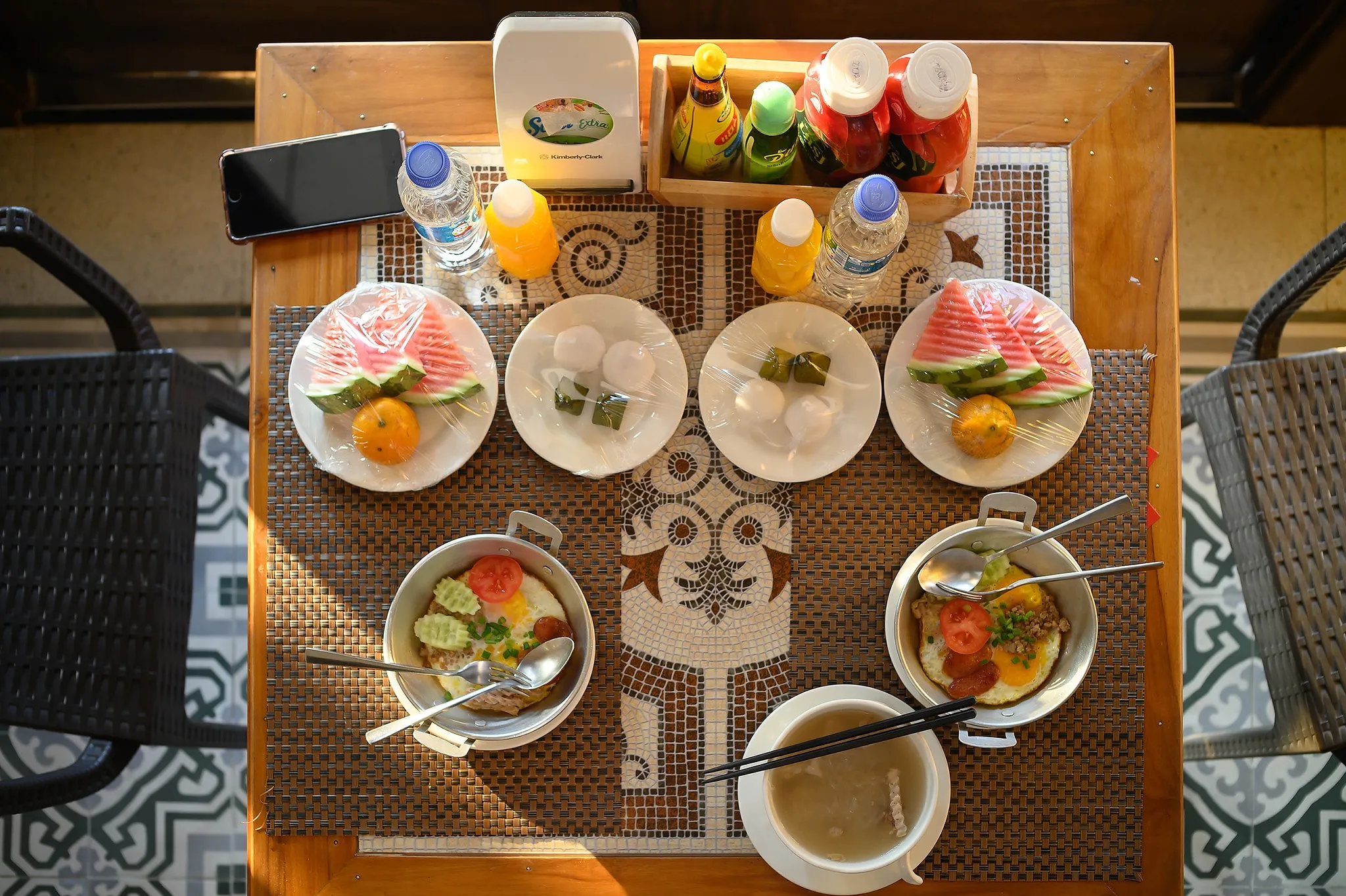 An overhead view of a breakfast table set for two with fruit plates, egg pans, and drinks