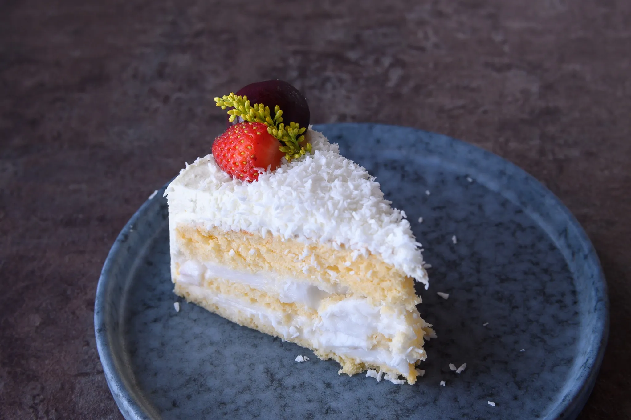 A slice of coconut layer cake topped with a strawberry on a dark stone plate
