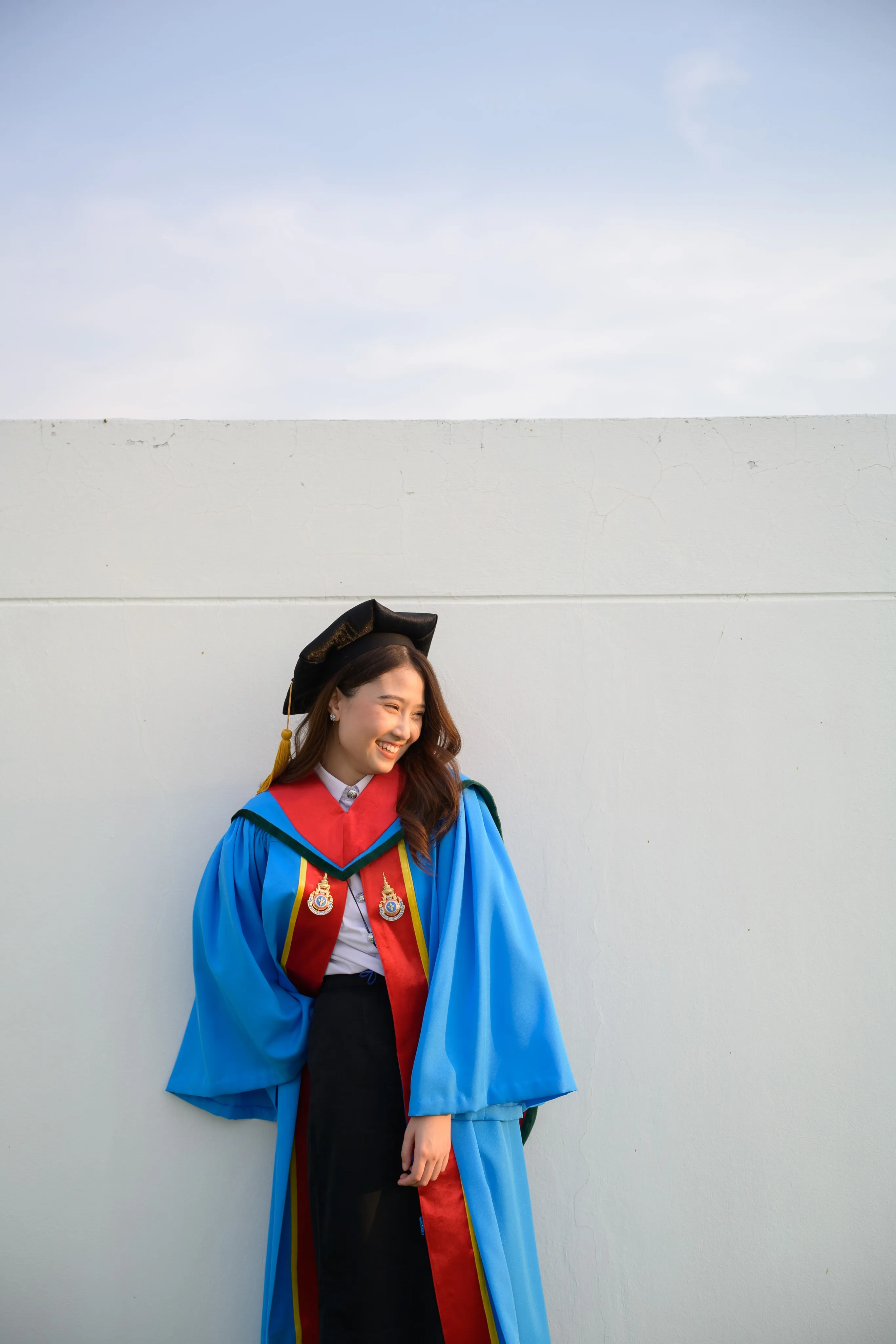 A laughing graduate in a mortarboard and blue gown leaning against a white wall