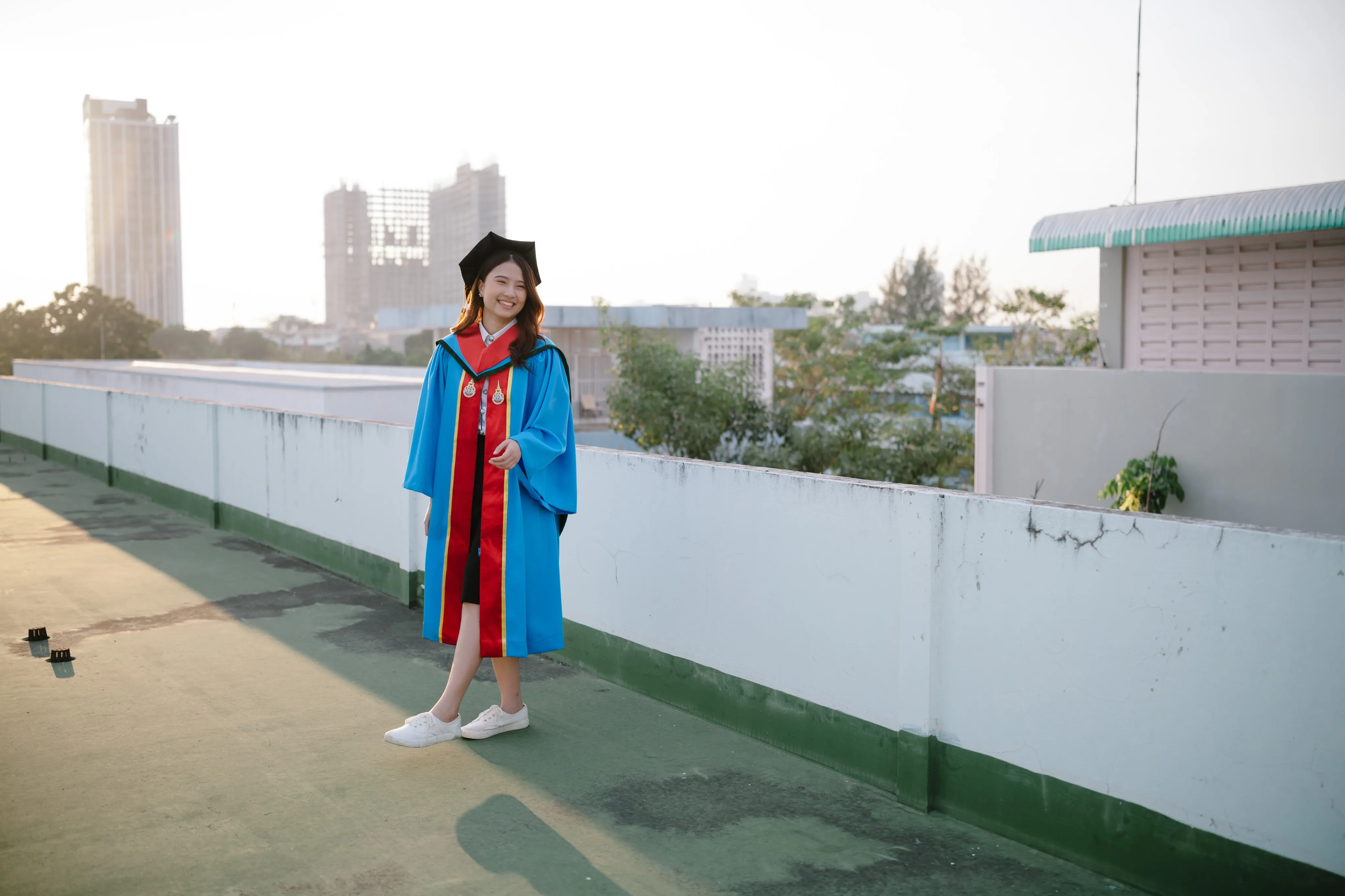 A graduate in a blue gown standing on a rooftop with the city skyline behind her