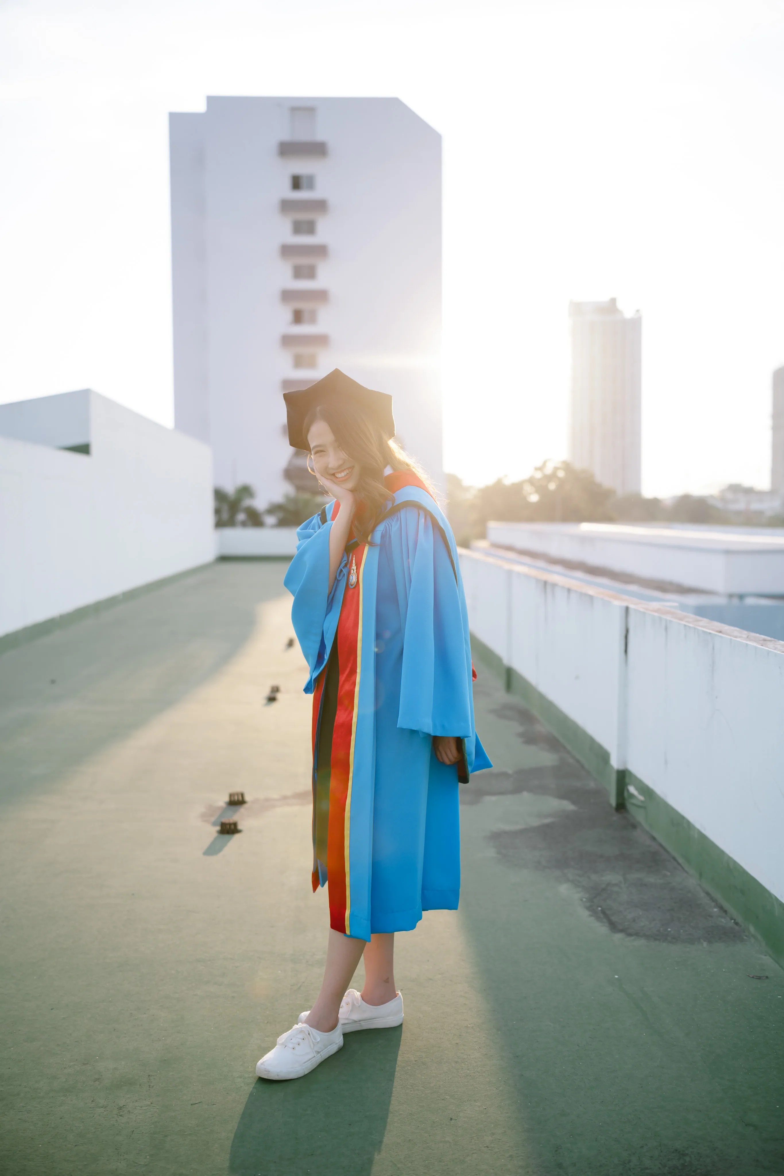 A graduate in a blue gown walking along a rooftop toward the camera, backlit by the sun