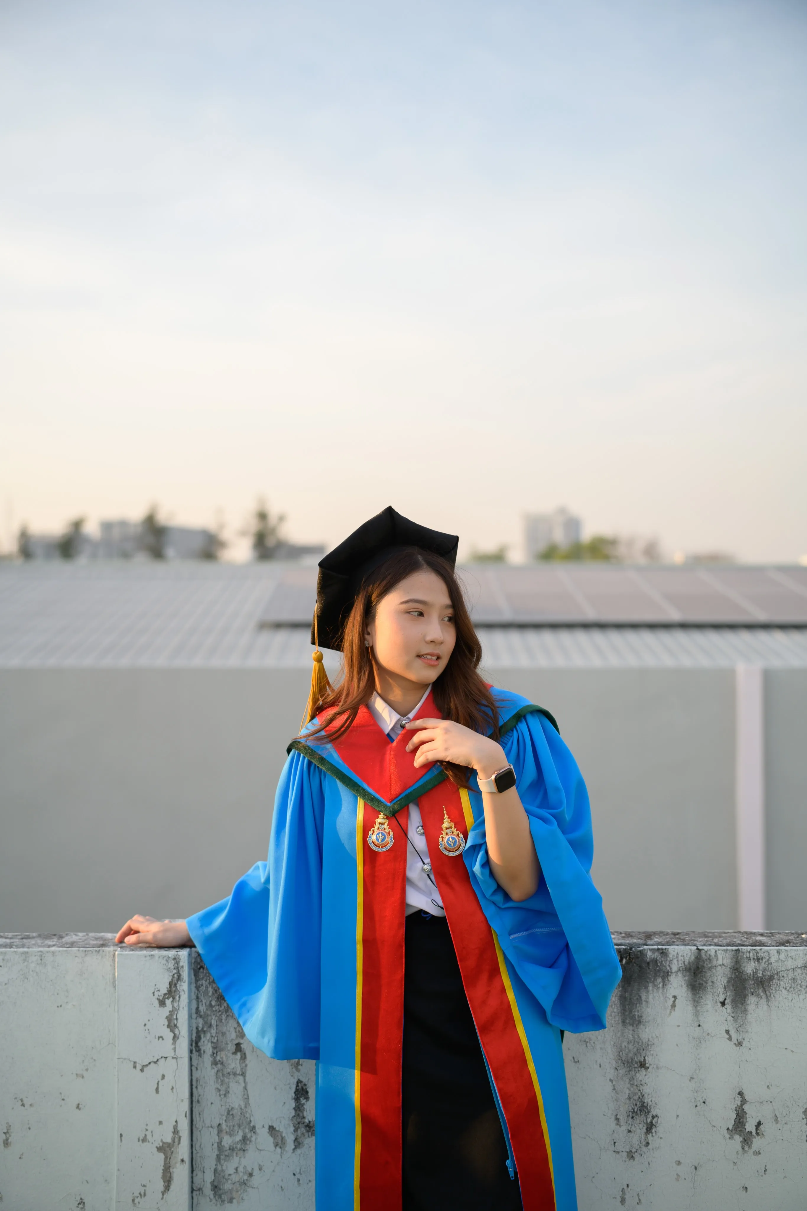 A graduate in a mortarboard and blue gown leaning on a rooftop ledge at sunset