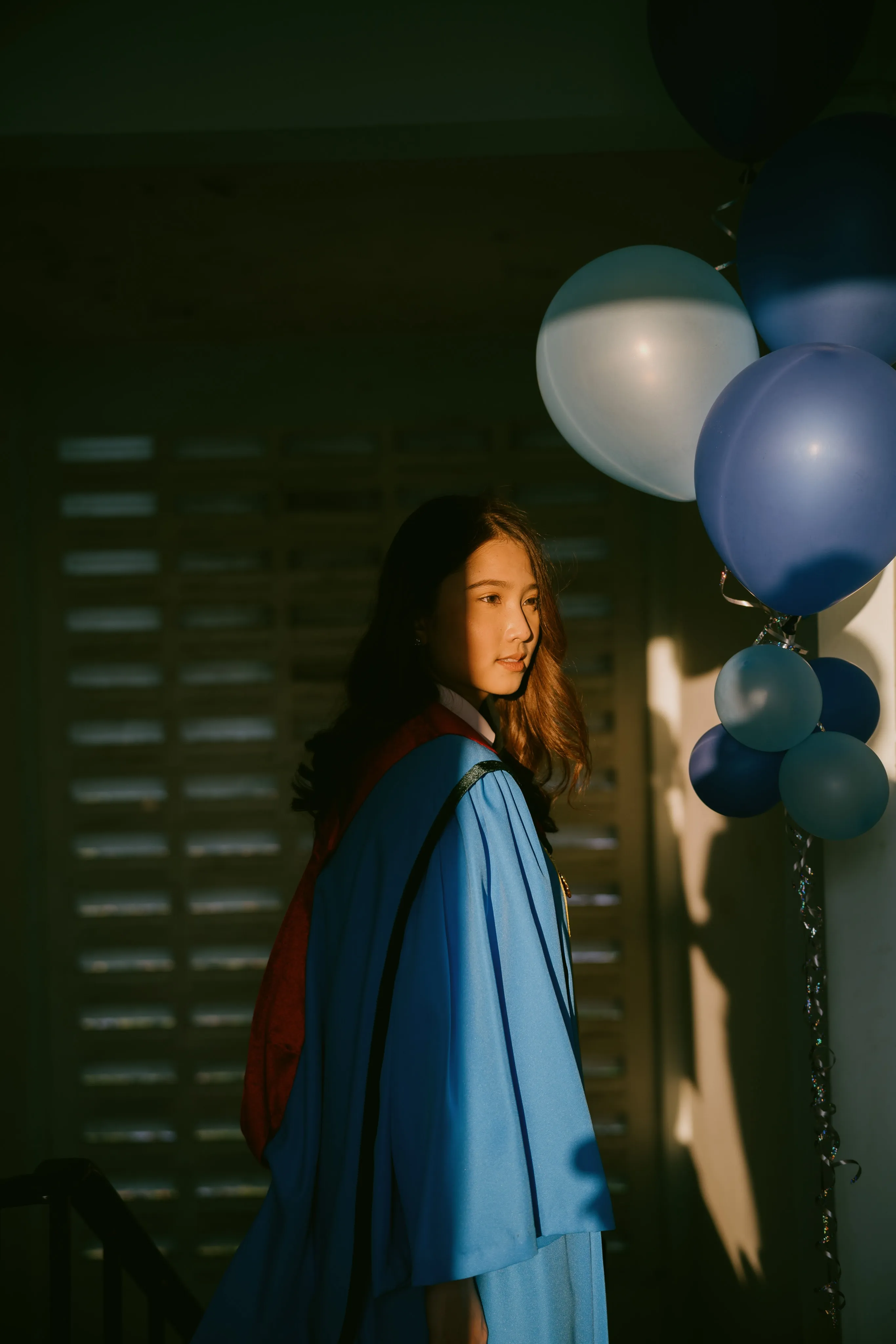 A graduate in a blue gown looking back in dim light beside a bunch of balloons