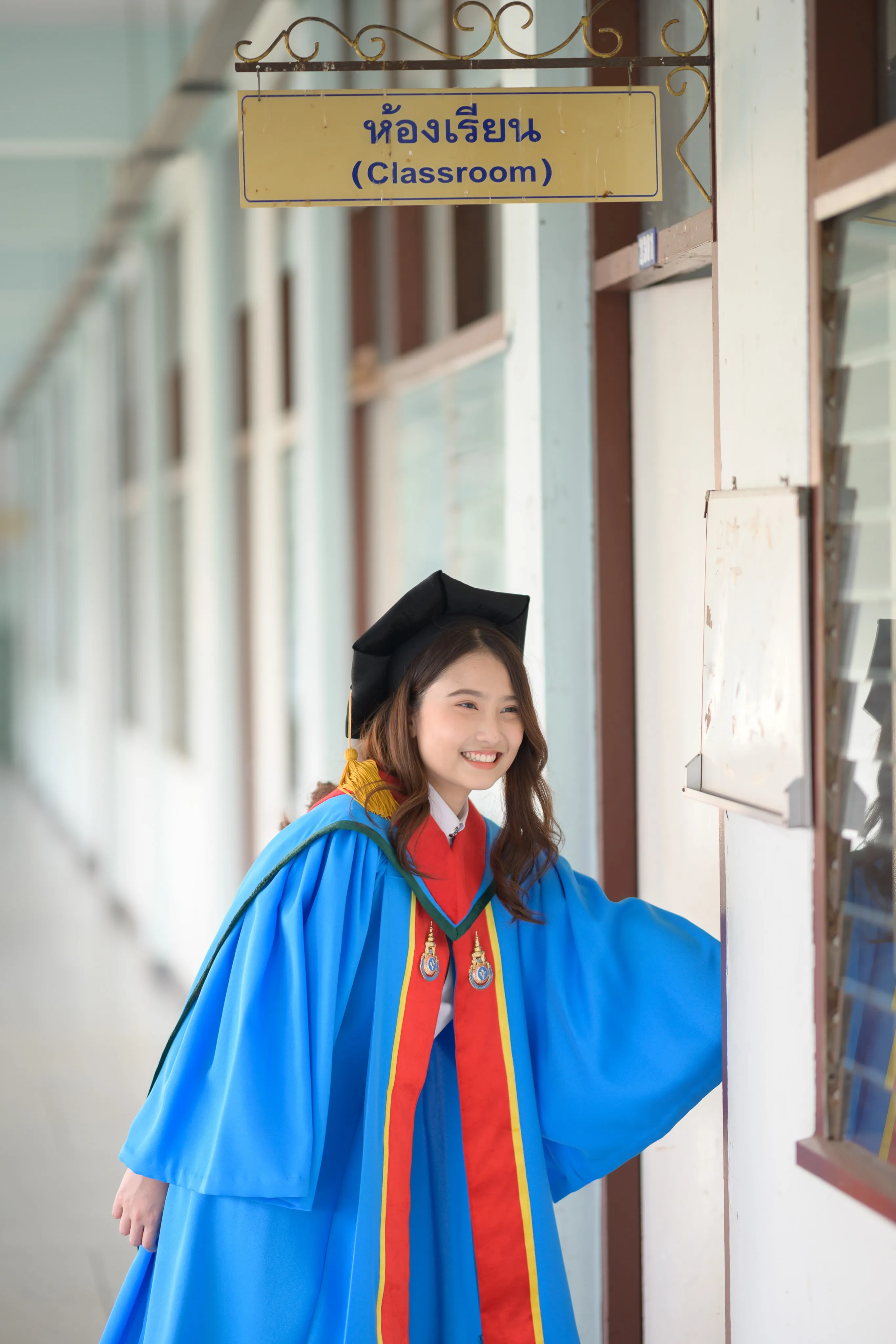 A smiling graduate in a mortarboard and blue gown leaning out of a classroom doorway