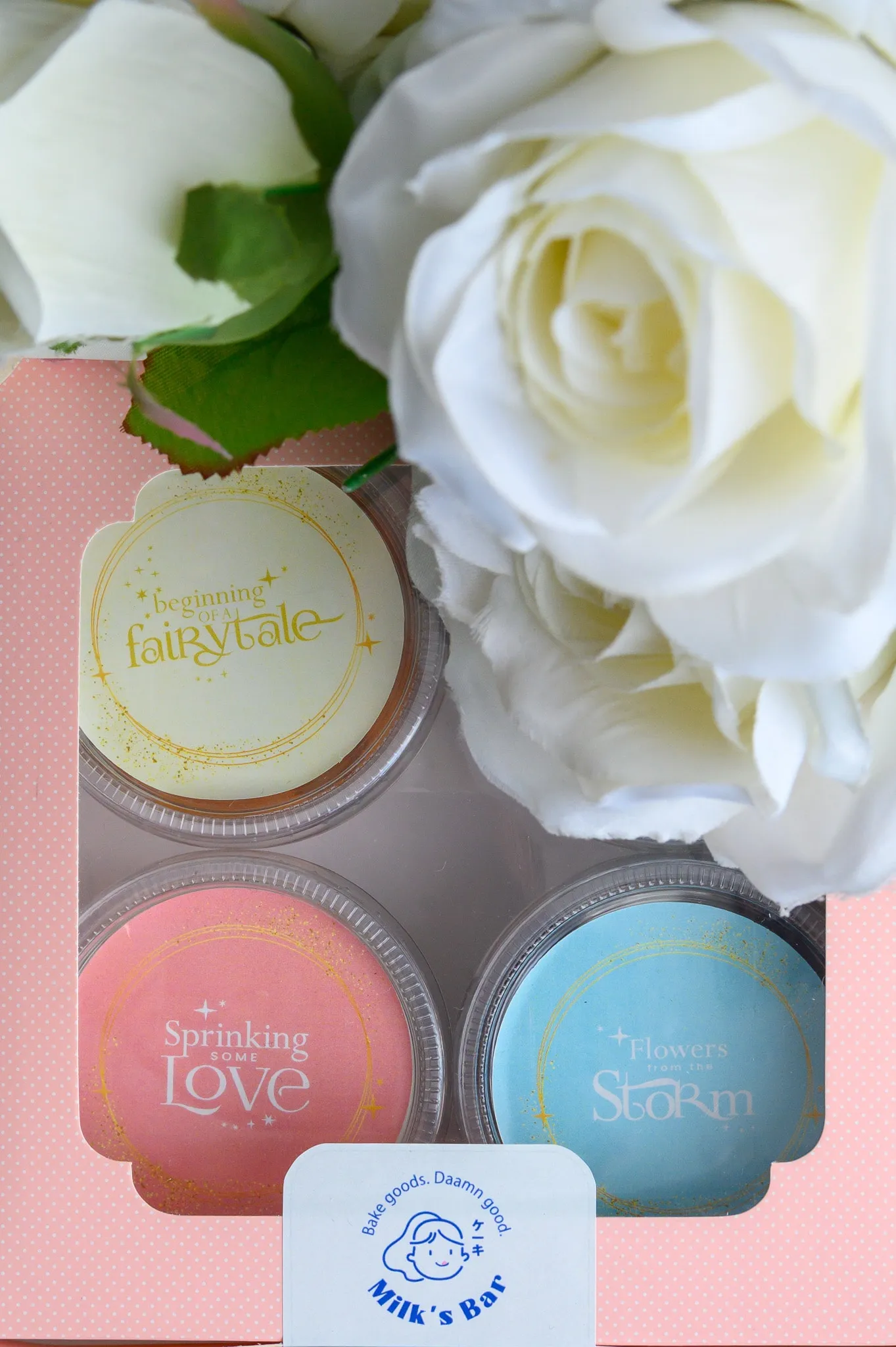 A gift box of four dessert cups with pastel labeled lids beside white roses