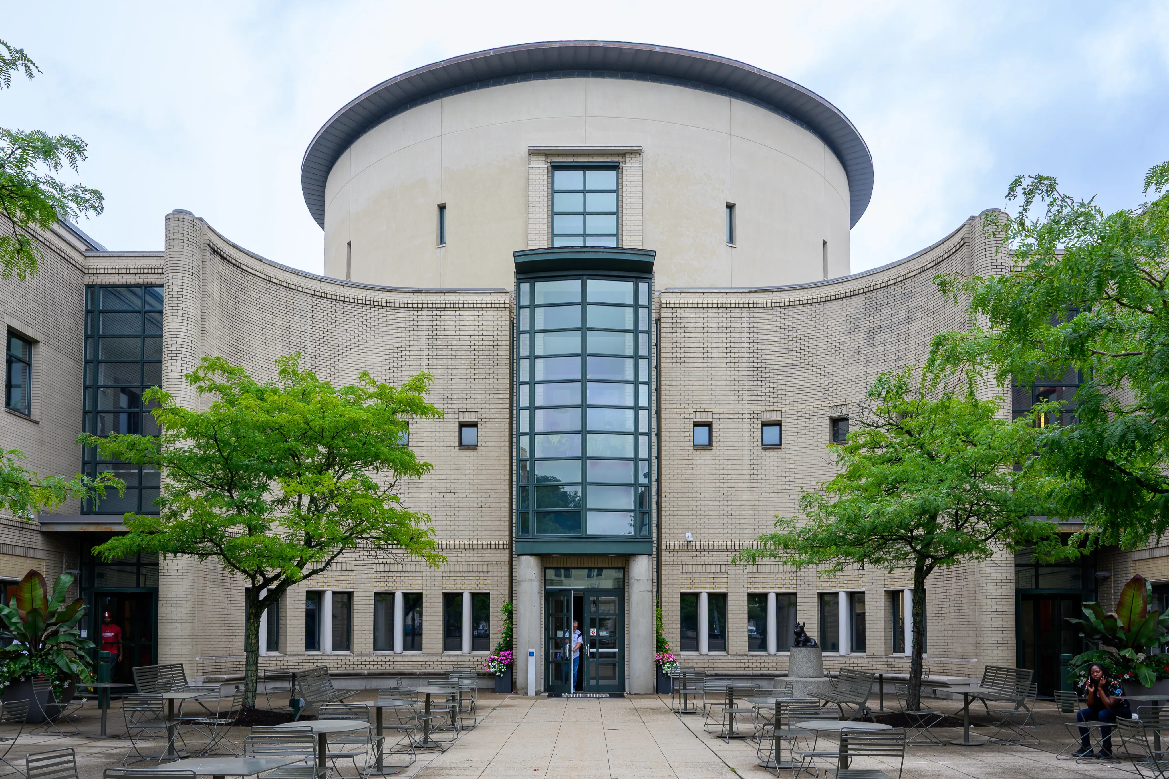 A curved tan brick facade with a round tower and a tall glass stair bay above the entrance, overlooking a courtyard of tables and chairs framed by leafy trees.